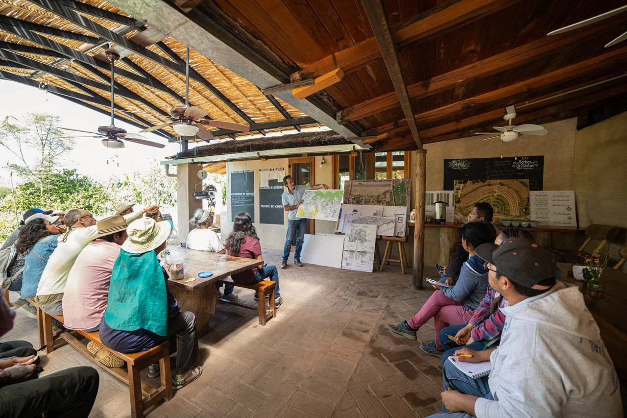 Clase o presentación al aire libre en un espacio cubierto de madera, con un grupo de personas observando a un instructor que muestra mapas y planos.