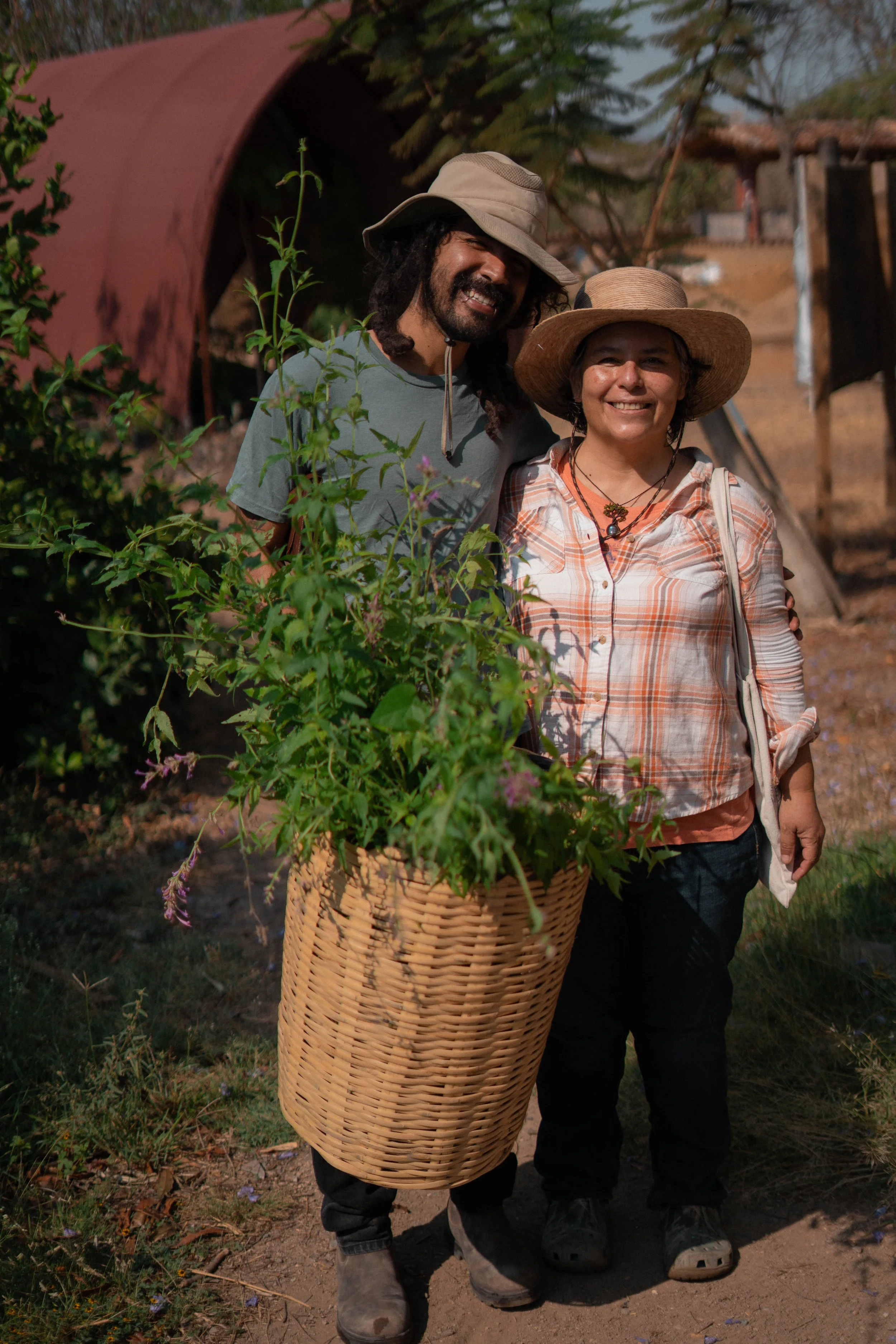 Dos personas, un hombre y una mujer, sonrientes, de pie en un campo, con un cesto grande de plantas con flores delante, usando sombreros de ala ancha, ropa de trabajo y botas, en un entorno rural con árboles y estructura de madera y barro al fondo.