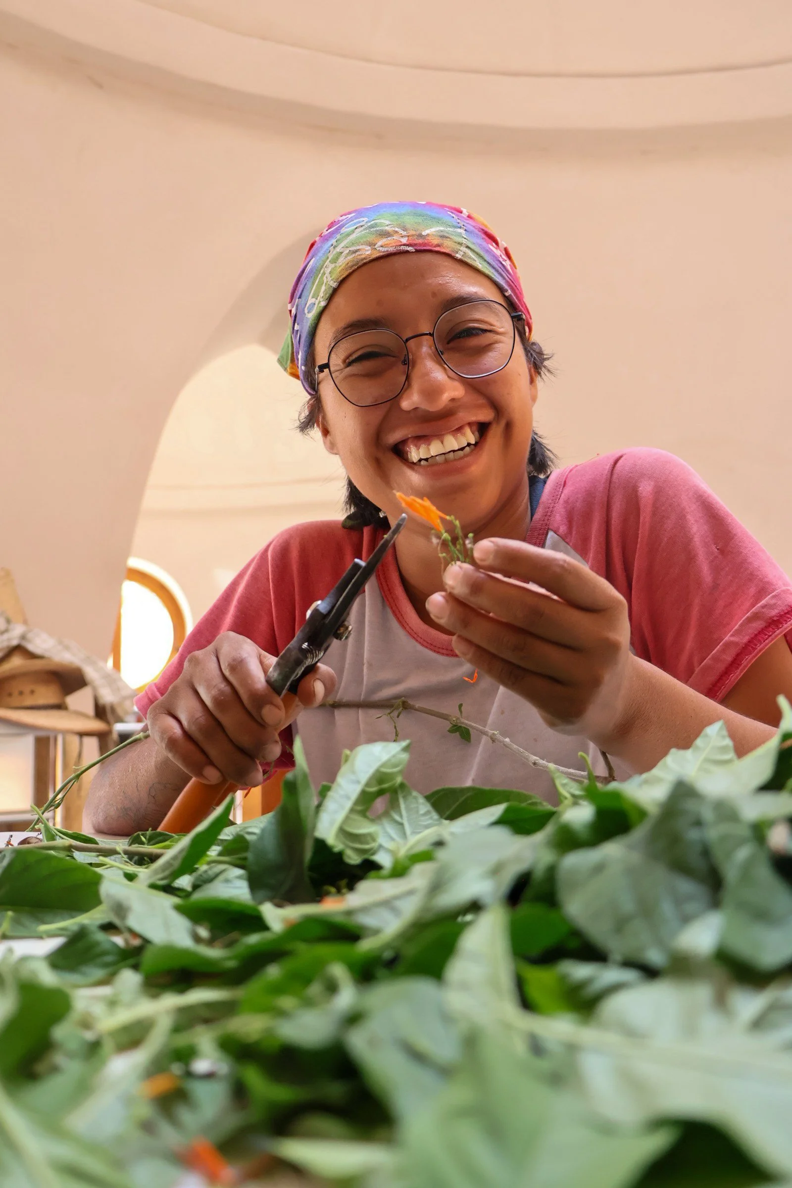 Persona sonriente con gafas y banda colorida en la cabeza sostiene unas tijeras y un pequeño ramo de flores en un entorno interior, rodeada de muchas hojas verdes.