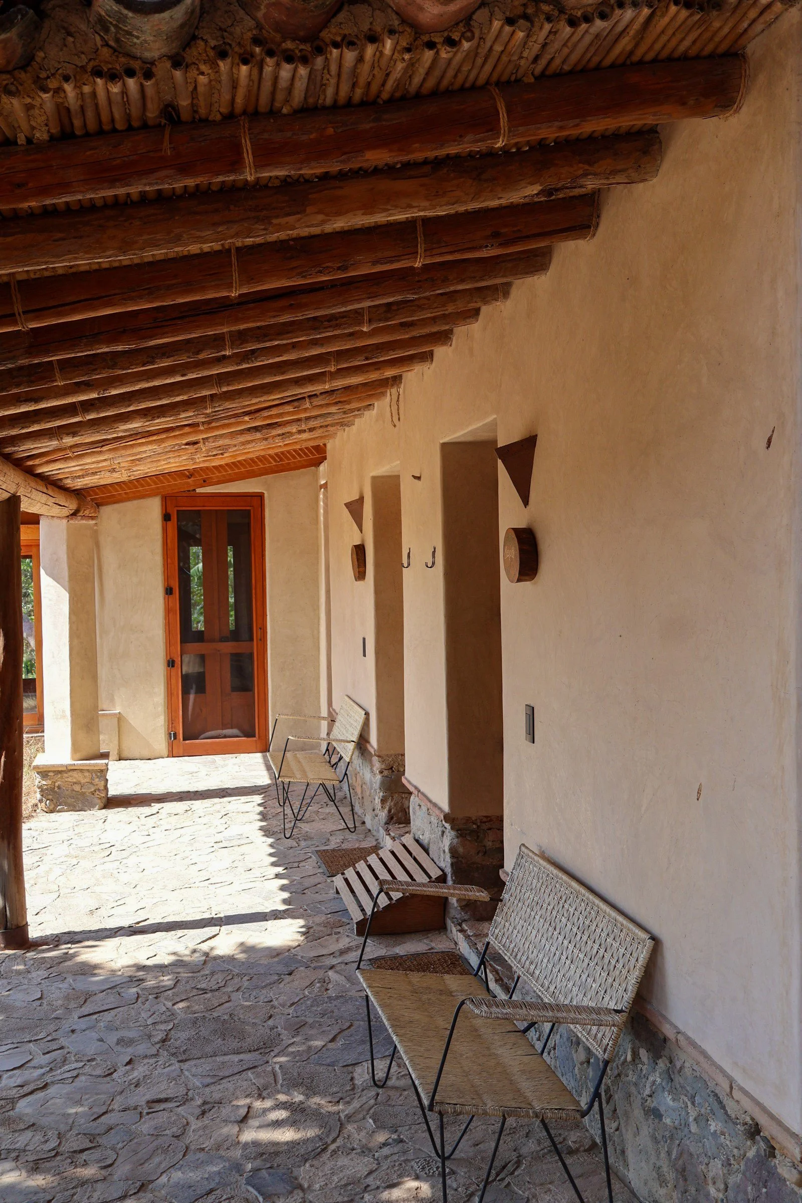 Un pasillo de estilo rústico con paredes de barro y techo de madera con vigas, decorado con varias sillas de mimbre y piedra en el piso.