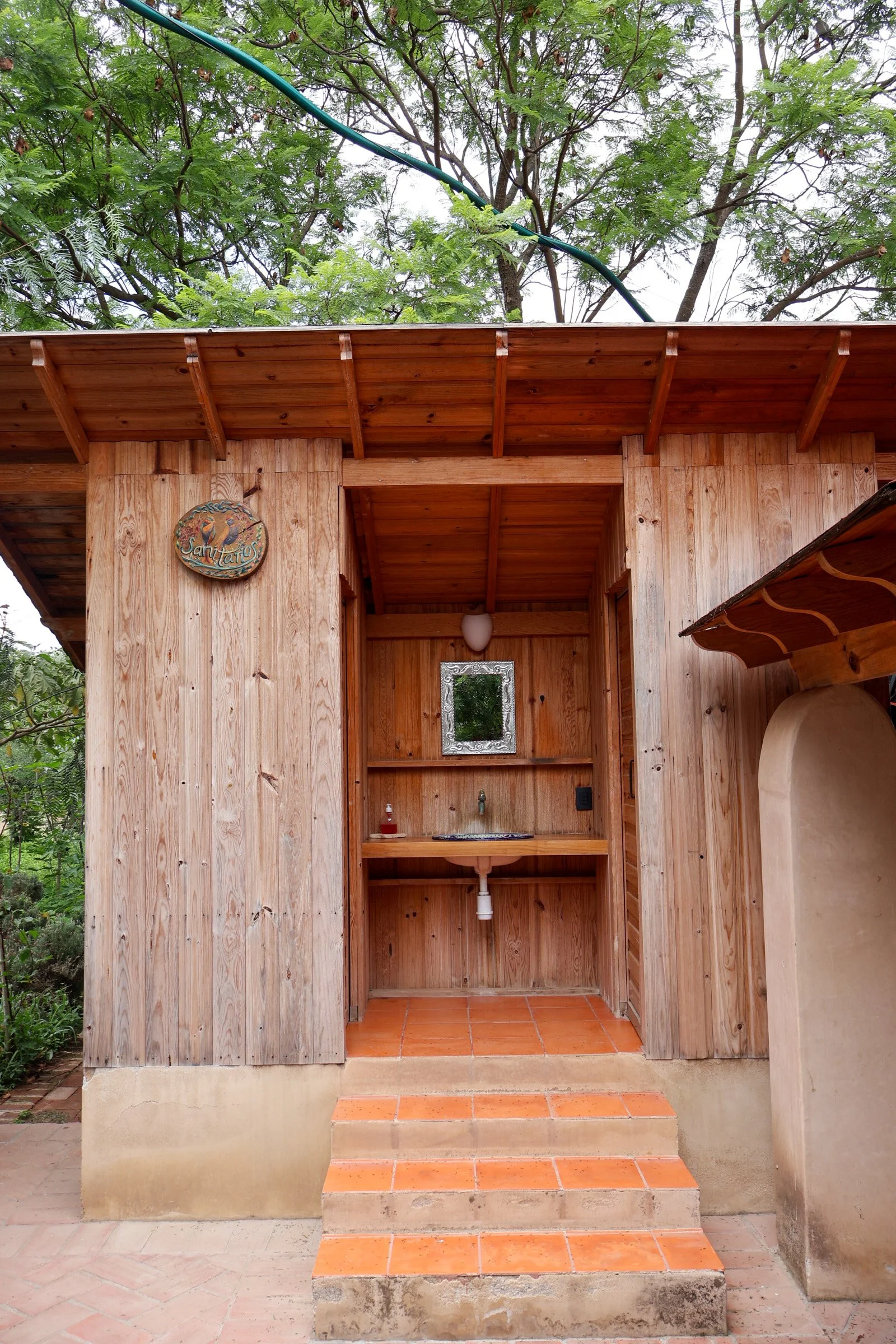 Estructura de madera con un lavabo y espejo, decorada con un signo que dice 'Sanitarios', rodeada de árboles verdes.