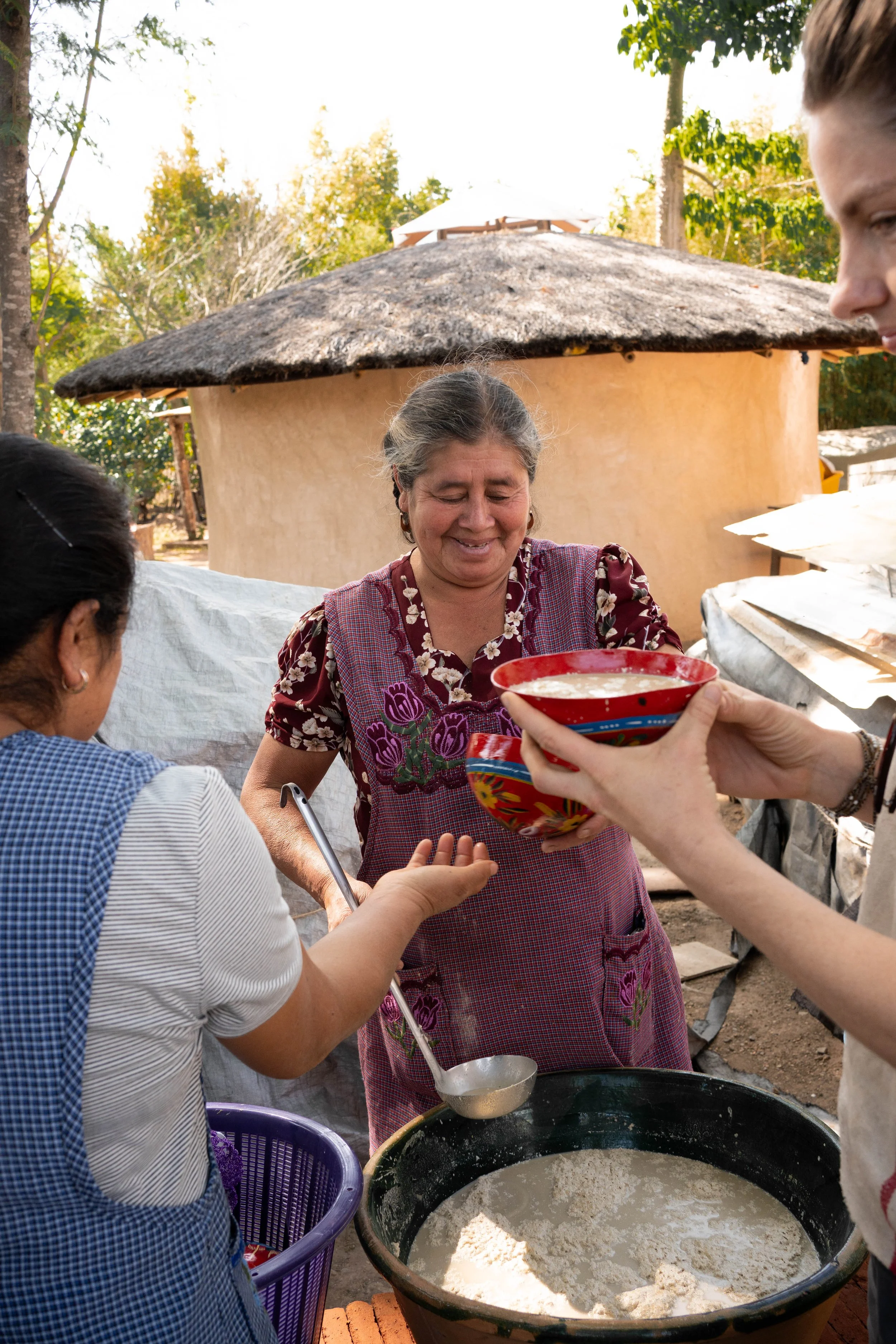 Mujer mayor sirviendo comida en un recipiente rojo a una mujer con una taza y un cuaderno, parte del esfuerzo por conseguir una alimentación sana en Tierra del Sol.
