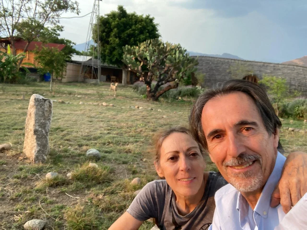 Una pareja toma una selfie en un campo abierto con árboles y rocas, en un entorno rural al atardecer, con un perro en el fondo y casas de fondo.