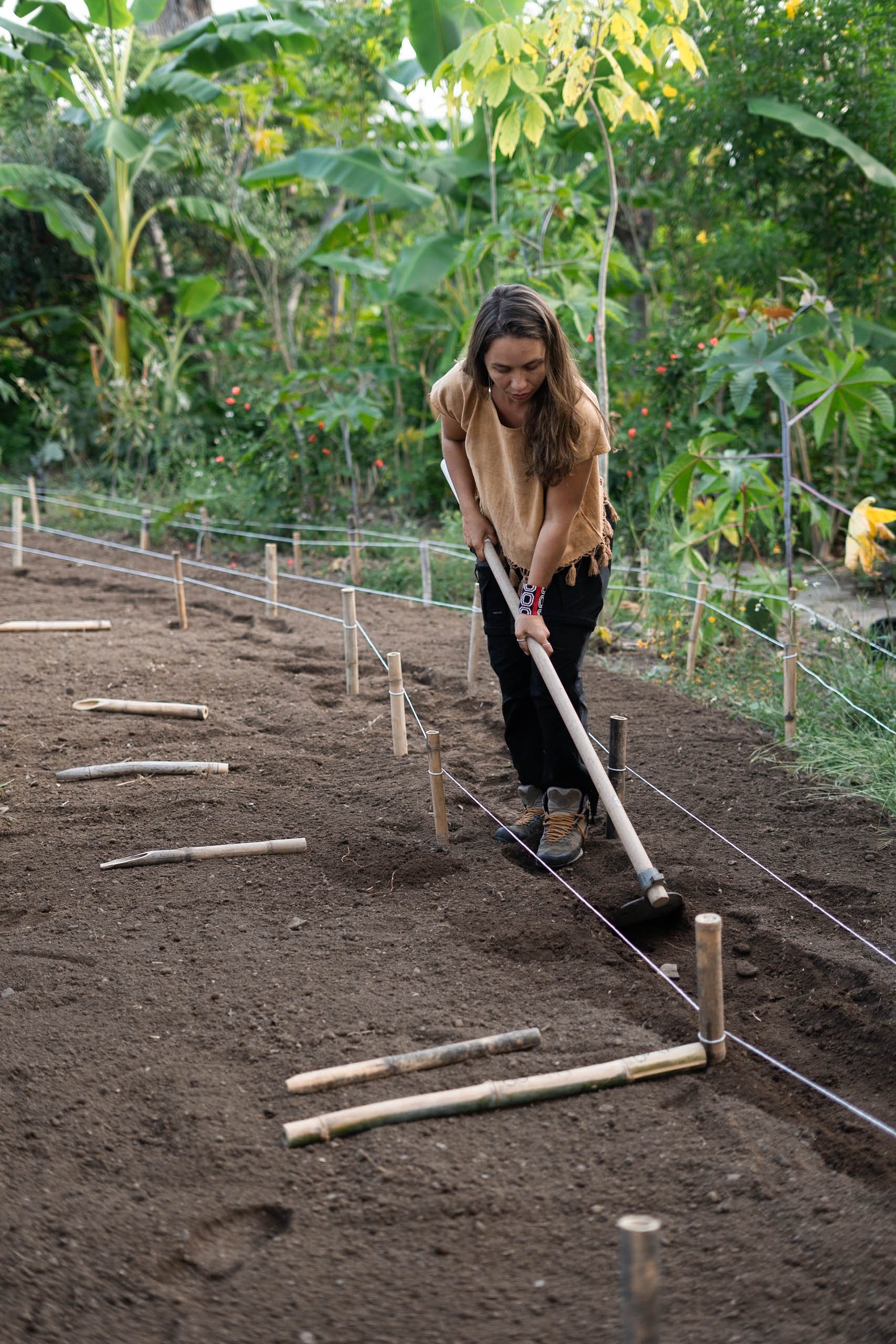 Mujer trabajando en un huerto, usando una herramienta para preparar la tierra, rodeada de plantas verdes y flores.