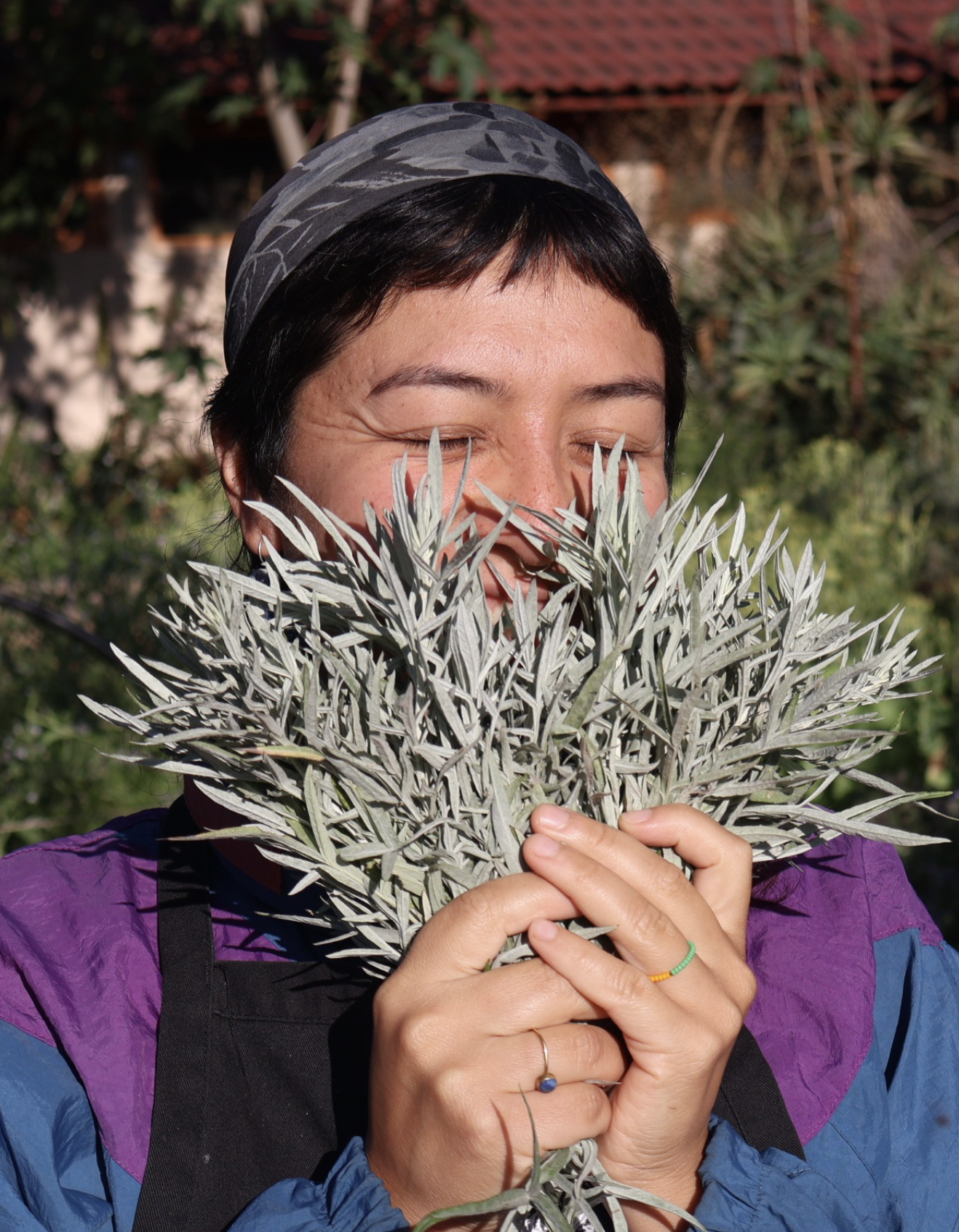 Una mujer sonriente sostiene un ramo de hojas plateadas en un entorno al aire libre, con un huerto sintrópico de fondo