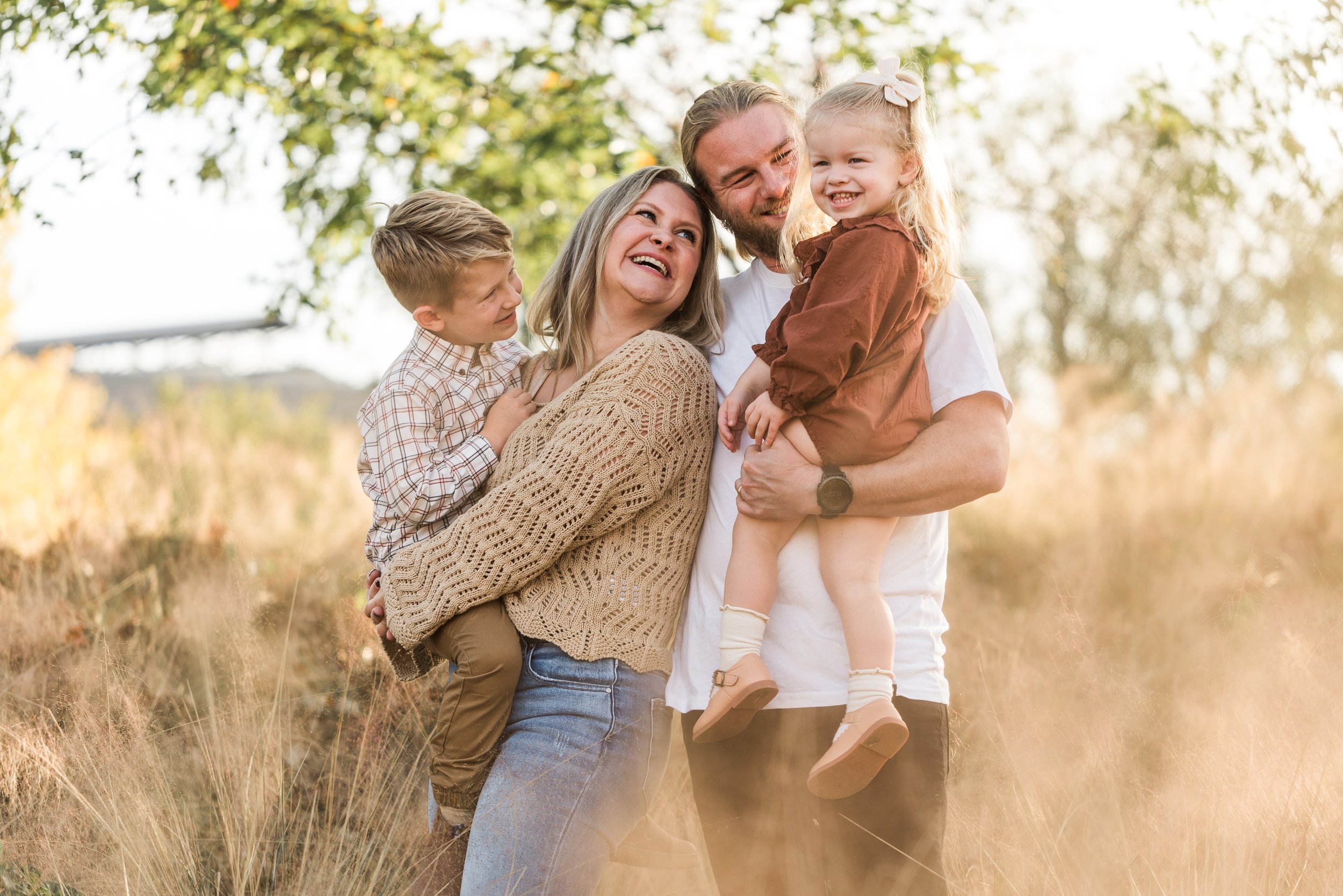 Railroad Park, Downtown Birmingham, AL - A happy family of four enjoying a moment outdoors. The mother, father, and two children are smiling and embracing, standing in a grassy field with trees in the background on a sunny day.