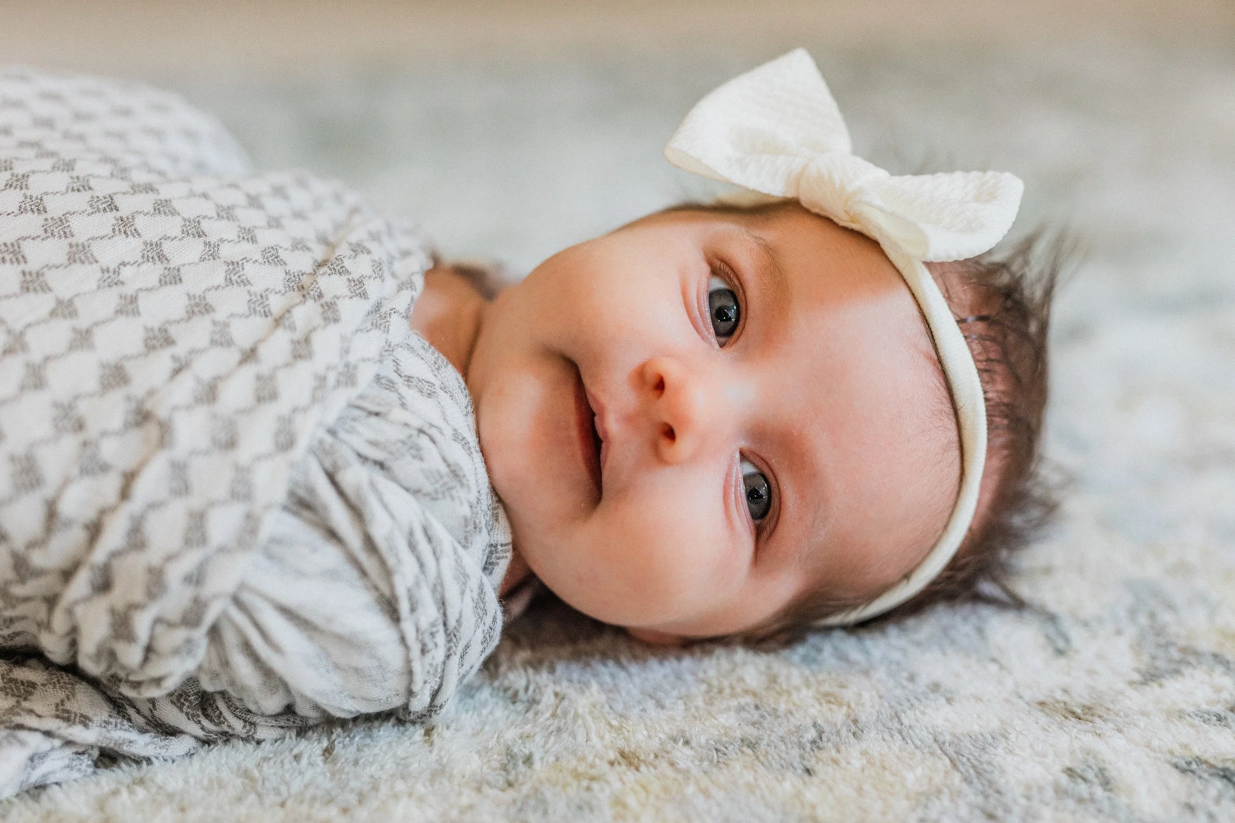 Inverness, AL - A baby girl swaddled wearing a white headband and bow, lying on a soft surface, smiling and looking at the camera.