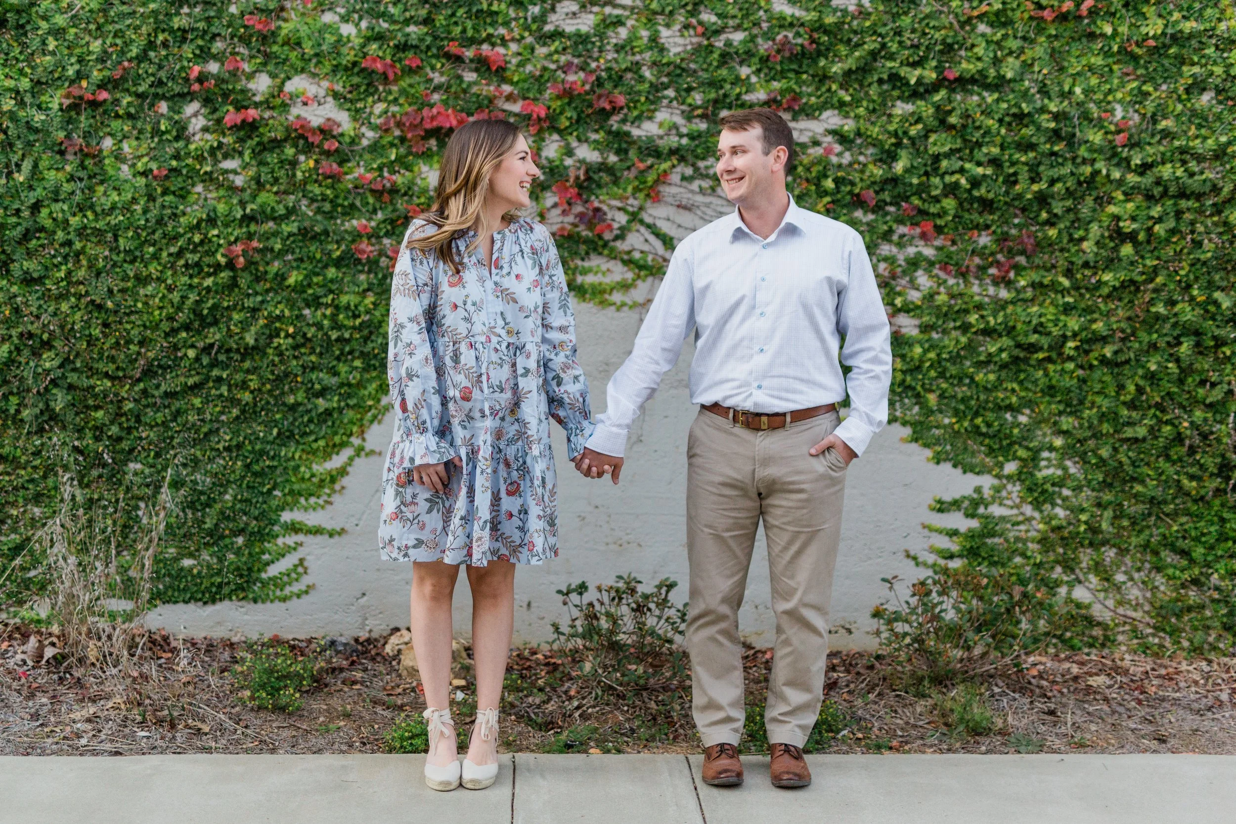 Railroad Park, Downtown Birmingham, AL - A man and woman holding hands, standing outside in front of a green leafy wall, smiling at each other.