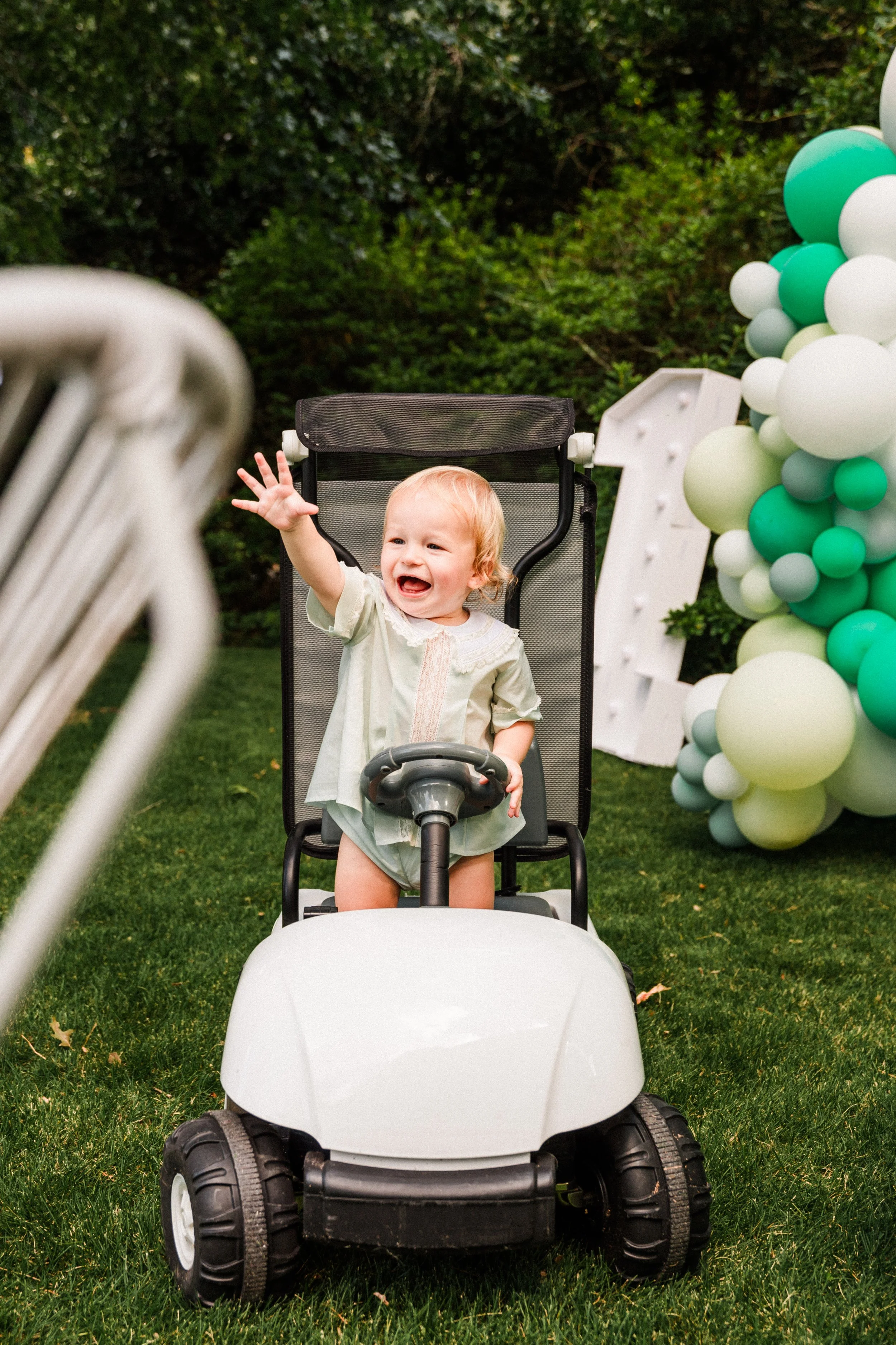 Mountain Brook, AL - A smiling young child riding a small vehicle at a celebration with balloons and a large letter '#1' decoration.