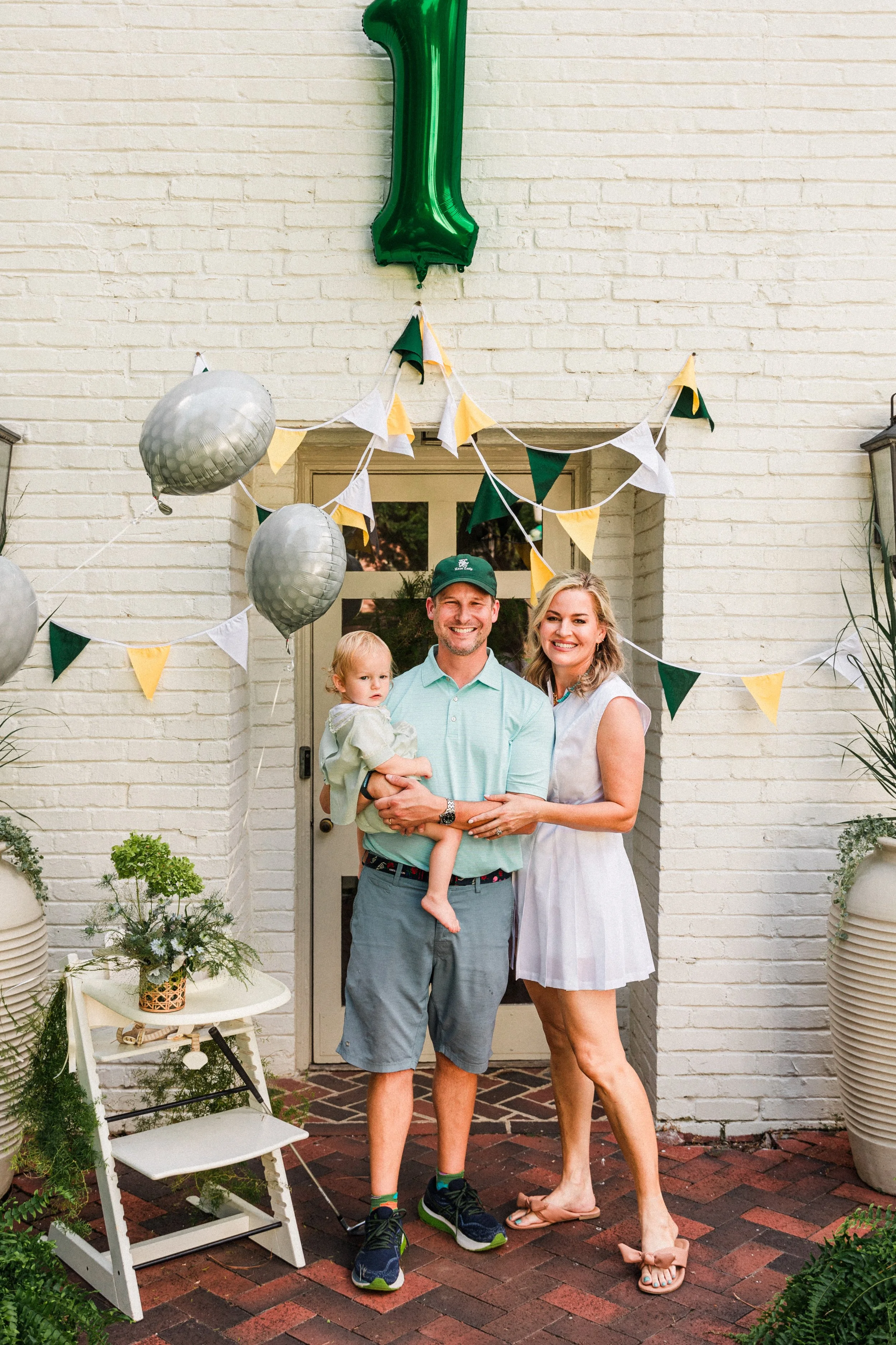 Mountain Brook, AL - A family of three celebrating a first birthday outside a house decorated with green, white, and gold balloons and bunting.