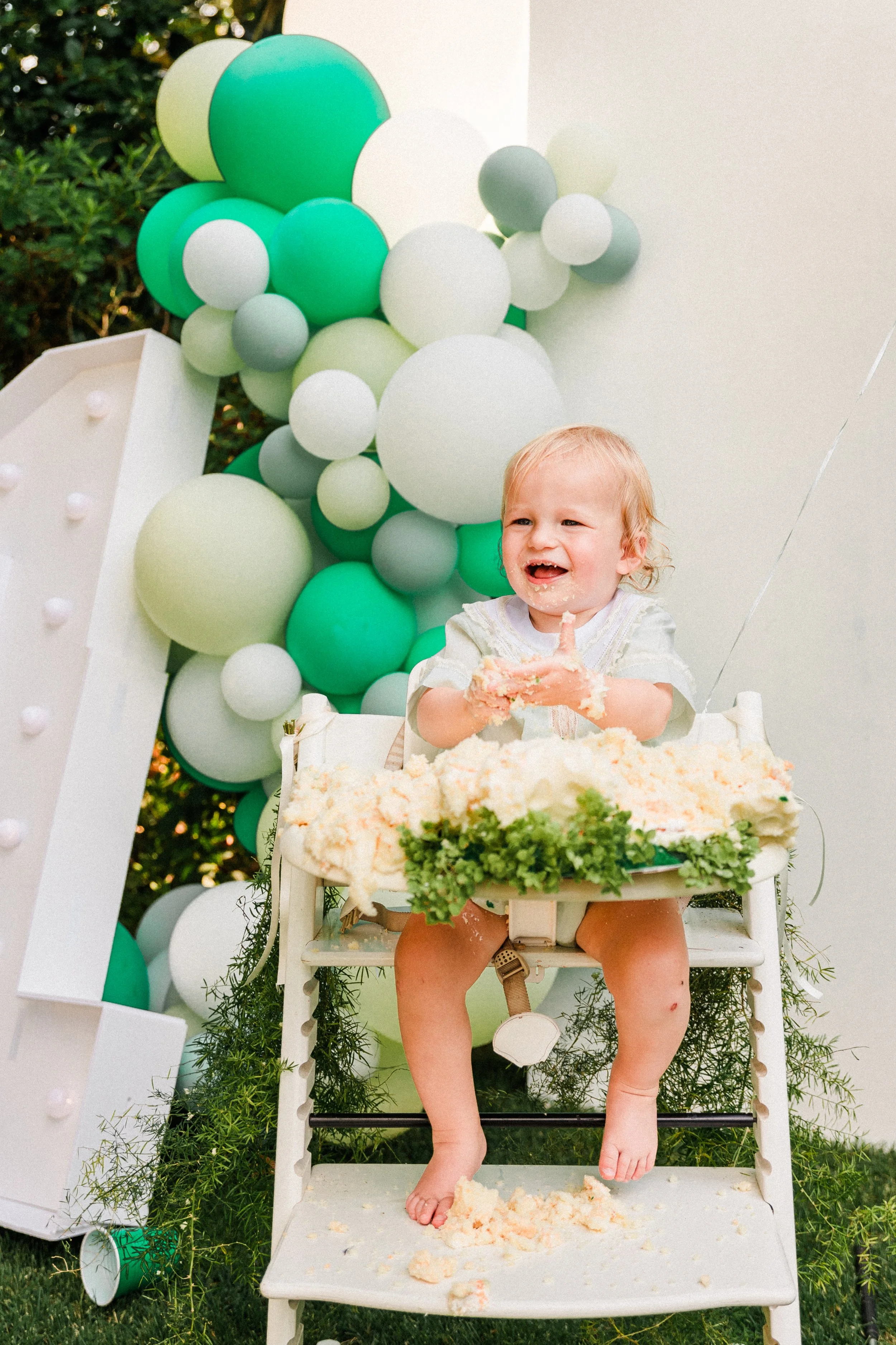 Mountain Brook, AL - A young child eating cake at a birthday celebration, sitting in a high chair with cake and cream on their face and hands, with a backdrop of green, white, and gray balloons and greenery.