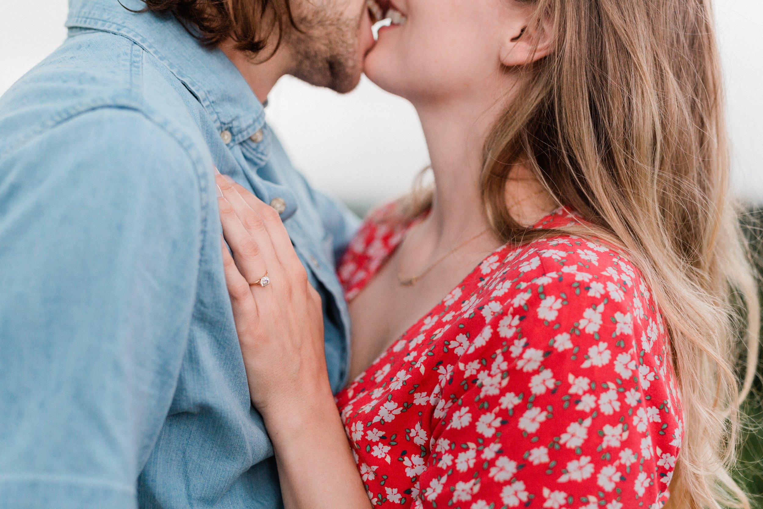 Vestavia, AL -A couple kissing, with the woman wearing a red dress with white floral pattern and an engagement ring, and the man in a light blue denim shirt.