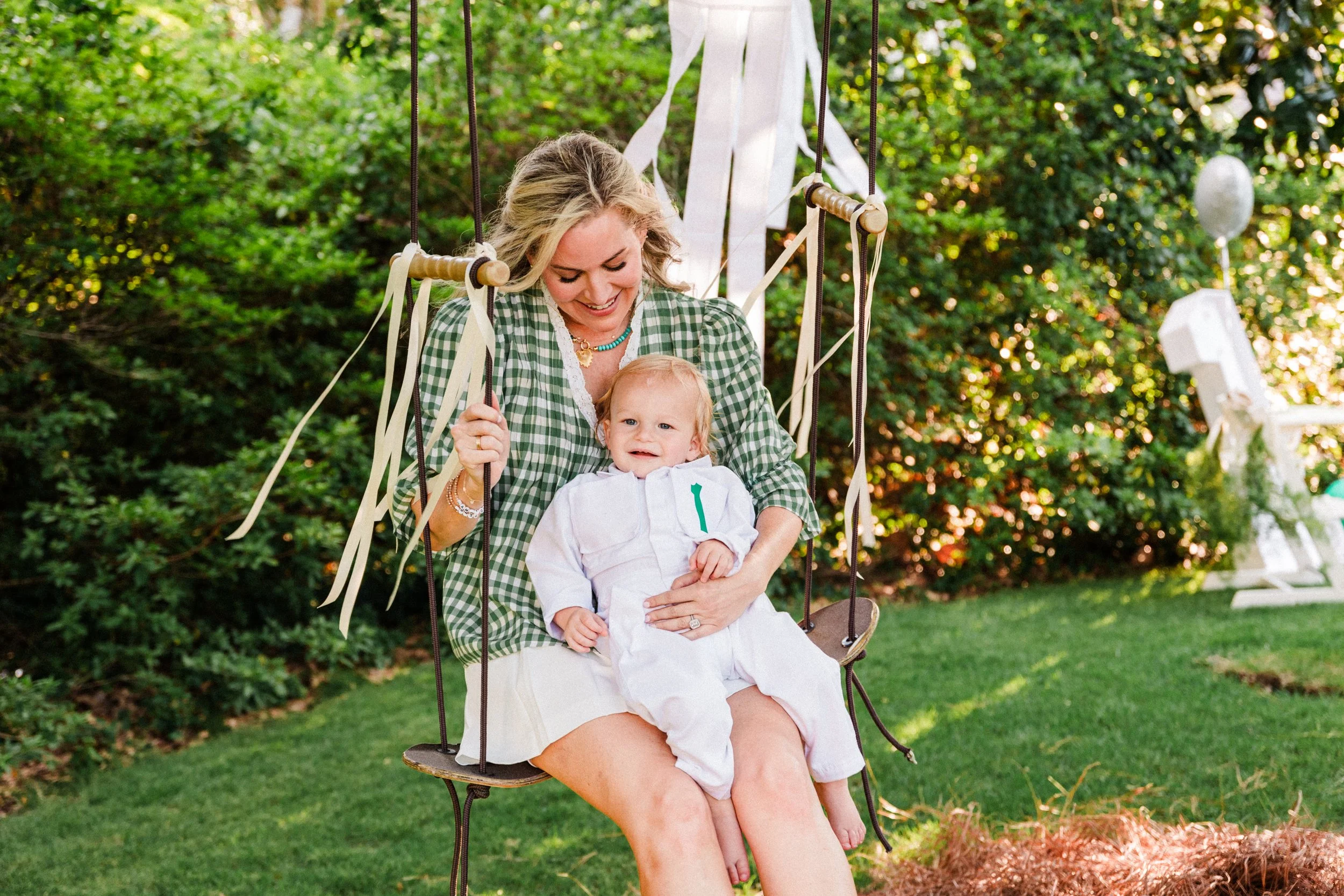 Mountain Brook, AL woman sits on a swing holding a young child on her lap, both are smiling. The woman wears a green checkered shirt and the child wears a white outfit. They are outdoors in a garden with trees and decorative chairs in the background.