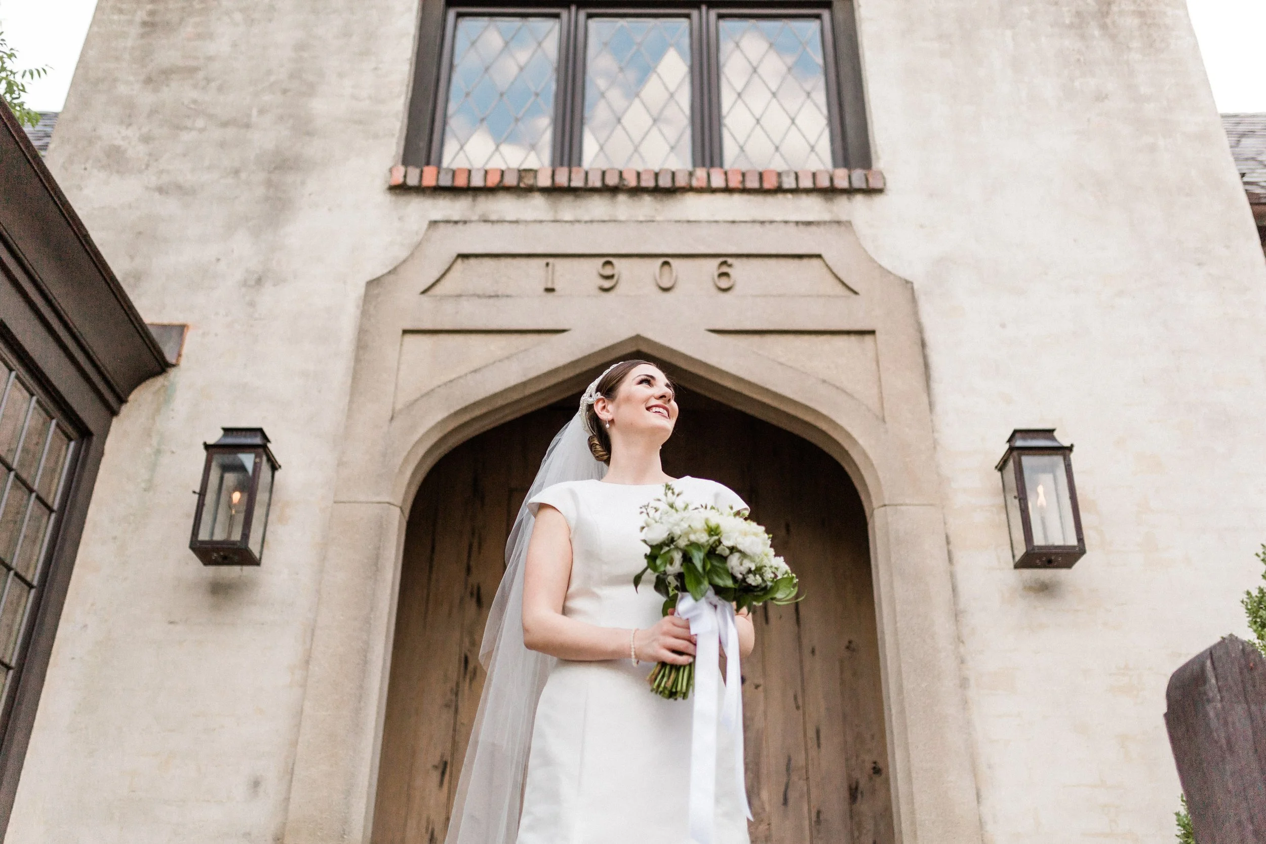 English Village, Mountain Brook, AL - A bride in a white wedding dress holding a bouquet of white flowers, standing in front of a church with the year 1906 inscribed above the entrance.