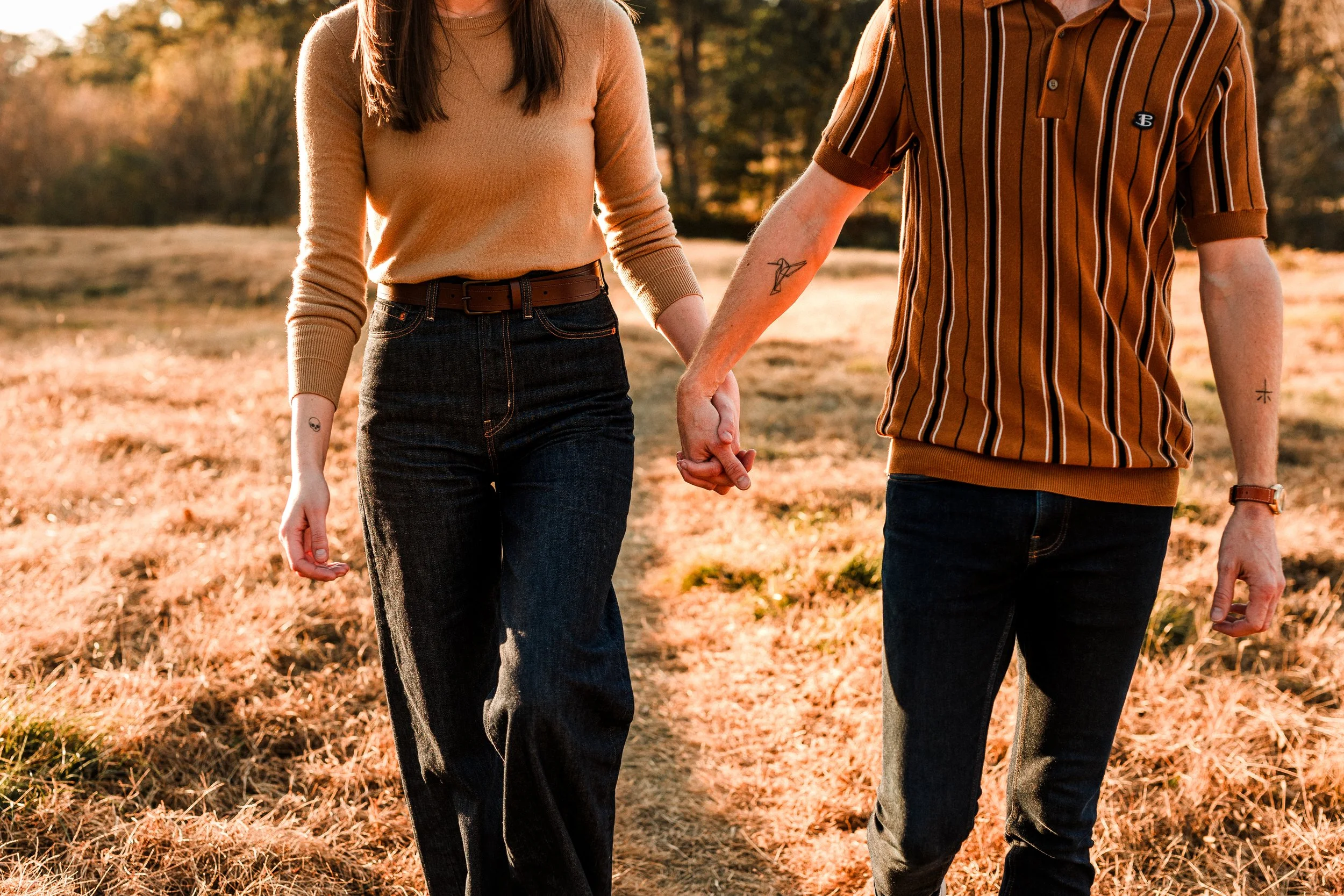 Vestavia, AL -A couple holding hands and walking outdoors in a grassy field during sunset, with trees in the background.