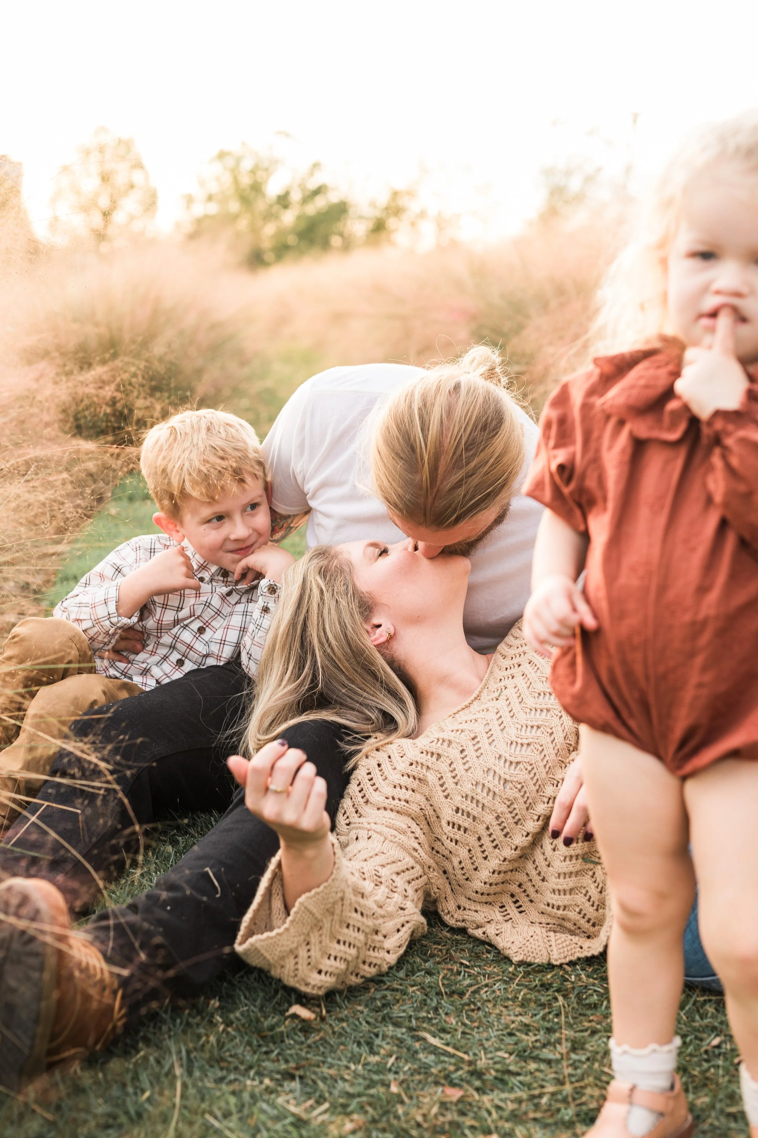 Railroad Park, Downtown Birmingham, AL - A family of four sharing a kiss and affection in a field of tall grass during sunset, with the mother lying on the ground and the father leaning over her, two children nearby.