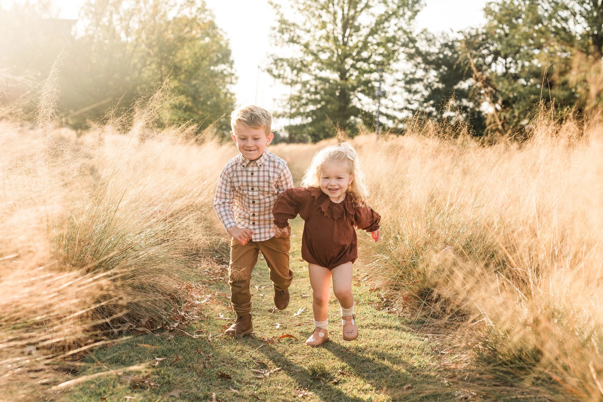 Railroad Park, Downtown Birmingham, AL - Two young children, a boy and a girl, running and smiling through a grassy field during sunset, holding hands.