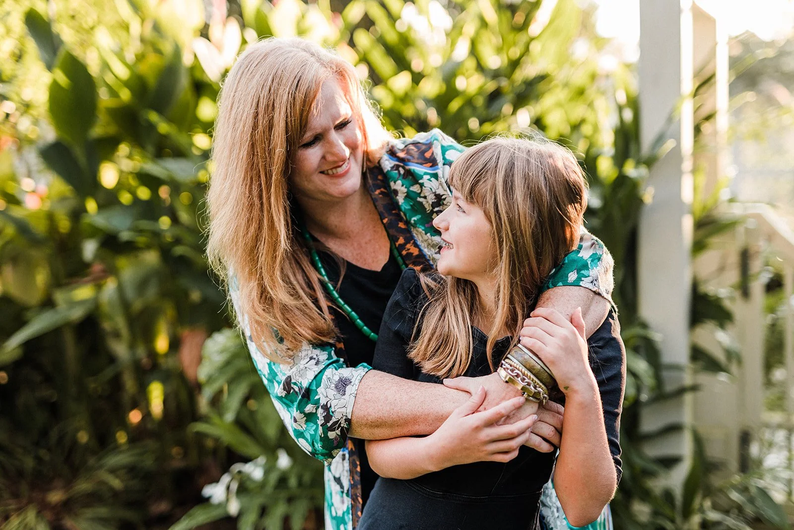 Birmingham Botanical Gardens - A woman and a young girl are smiling and sharing a moment of affection outdoors among tall, green plants. The woman has long red hair and is wearing a floral patterned shirt with a black top, and the girl has long blond
