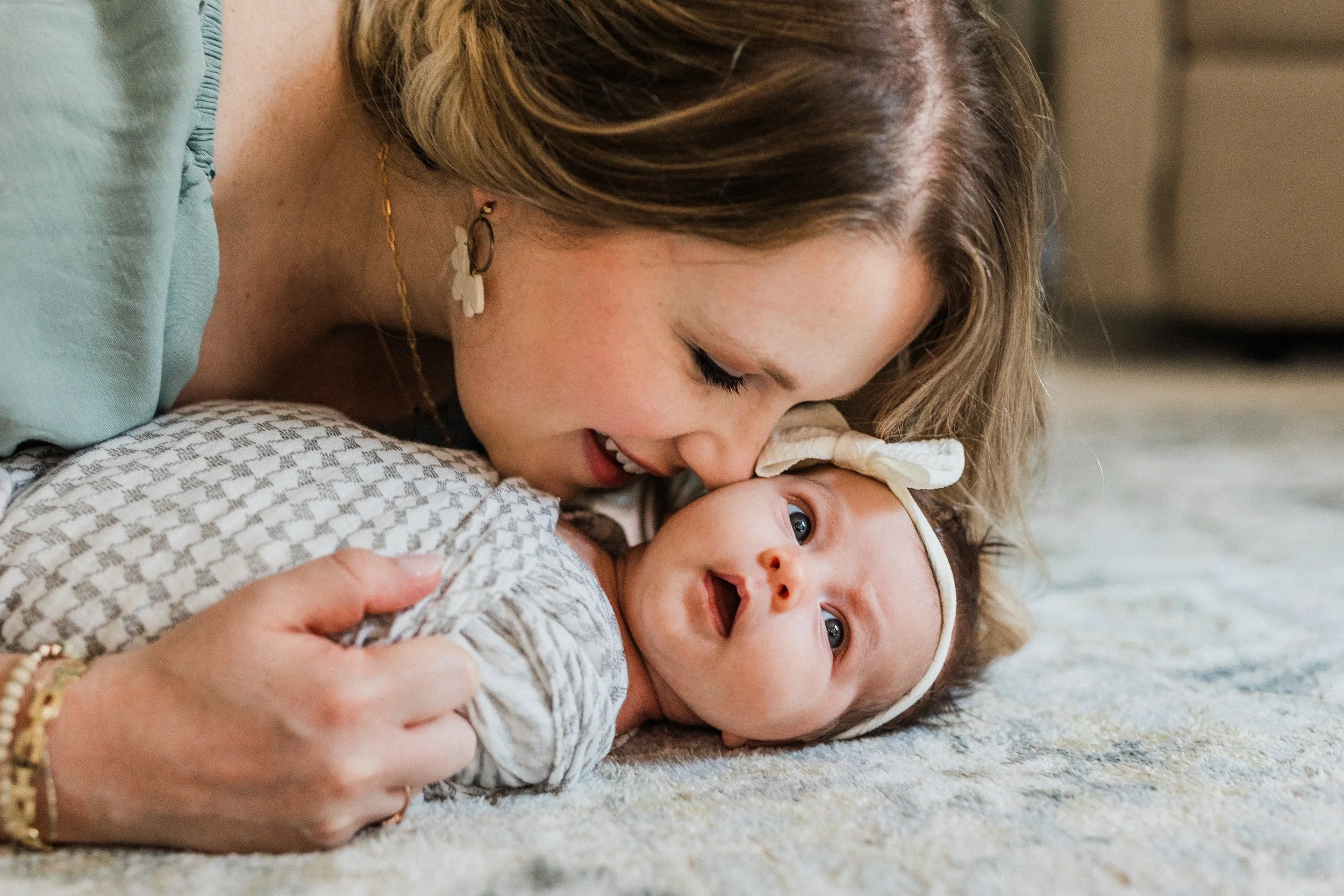 Inverness, AL - Woman is snuggling and kissing her newborn baby, who is looking at the camera with a headband and open mouth.