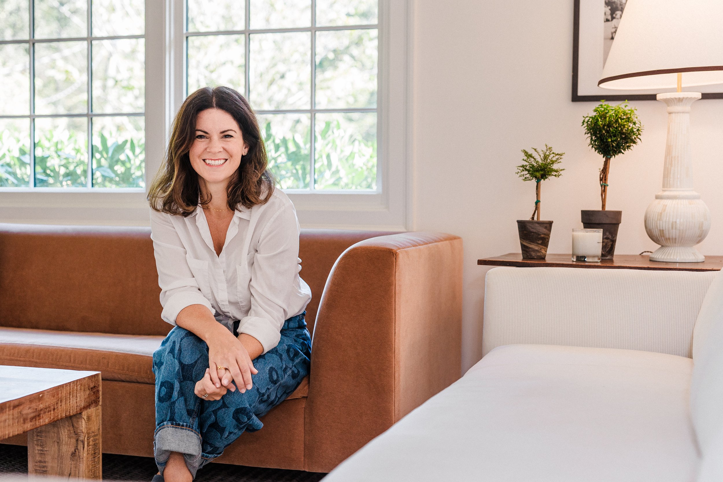 Cahaba Heights, AL - A woman with dark brown hair and a white shirt sitting on a tan couch in a bright living room with large windows, white walls, and decorative plants on a wooden table.