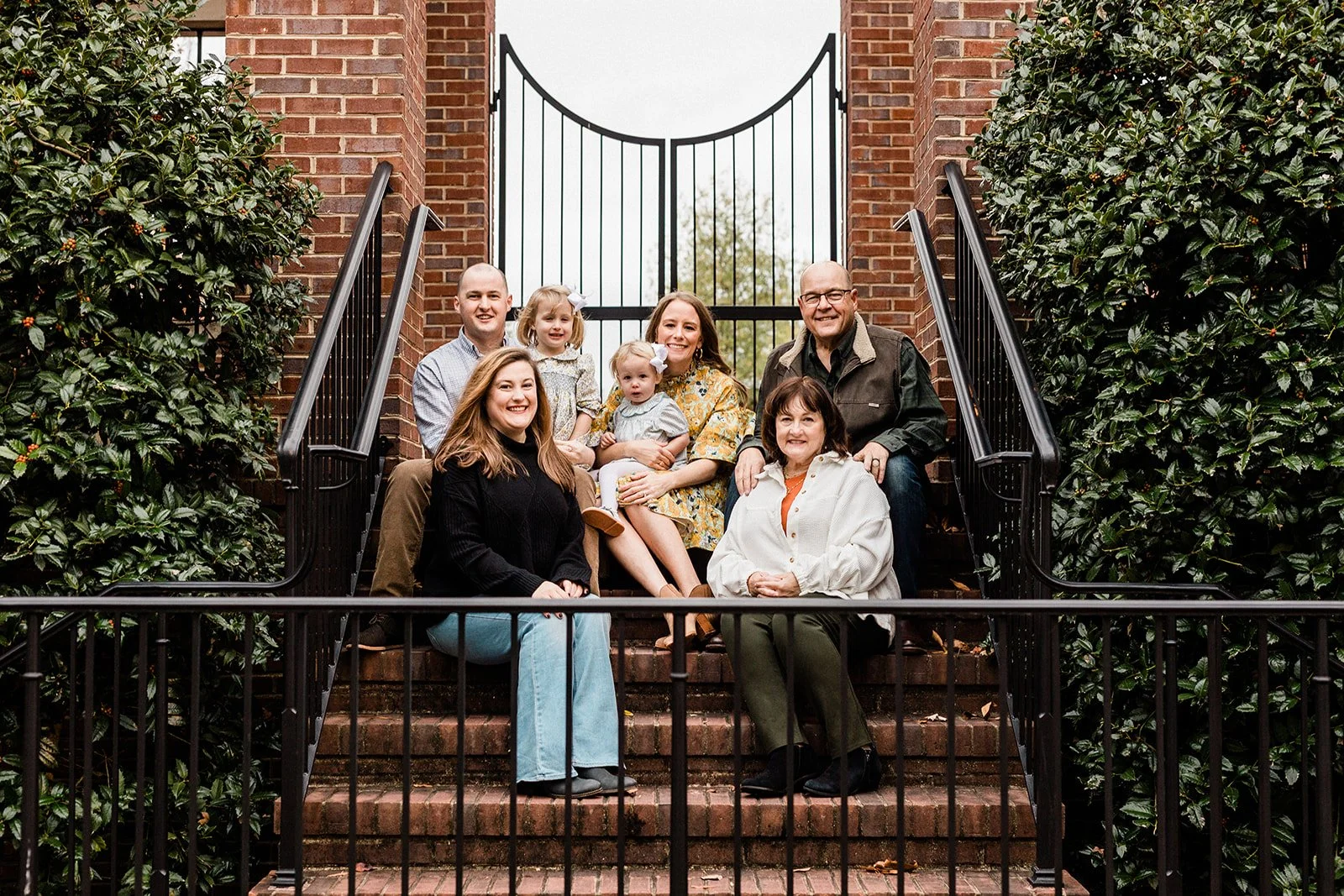 Moss Rock, Bluff Park, AL - An extended family of eight people, including adults and children, sitting on brick steps outside a building with a gated entrance, surrounded by bushes.