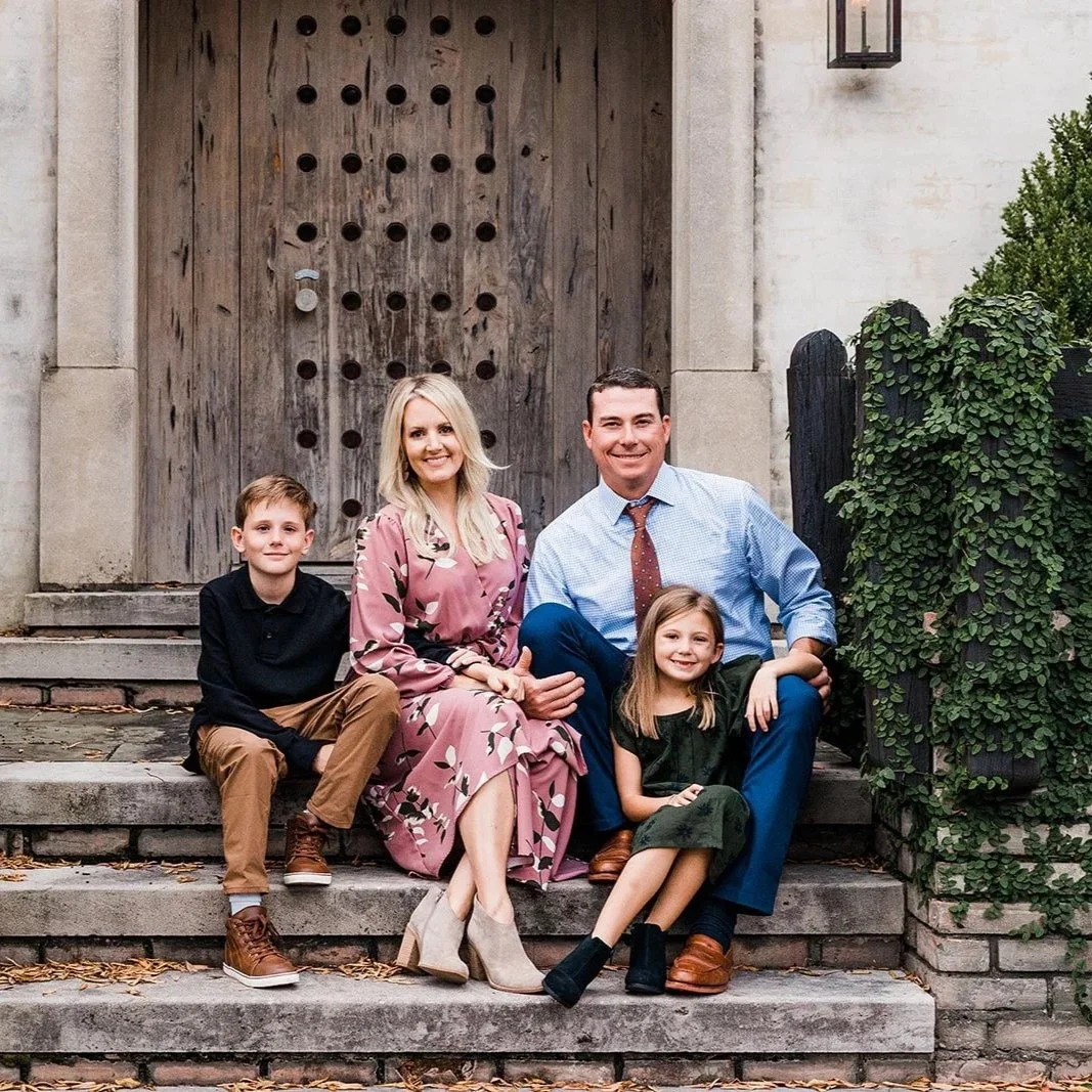 English Village, Mountain Brook, AL - A family of four sitting on outdoor stairs in front of a unique, large wooden door, smiling at the camera.