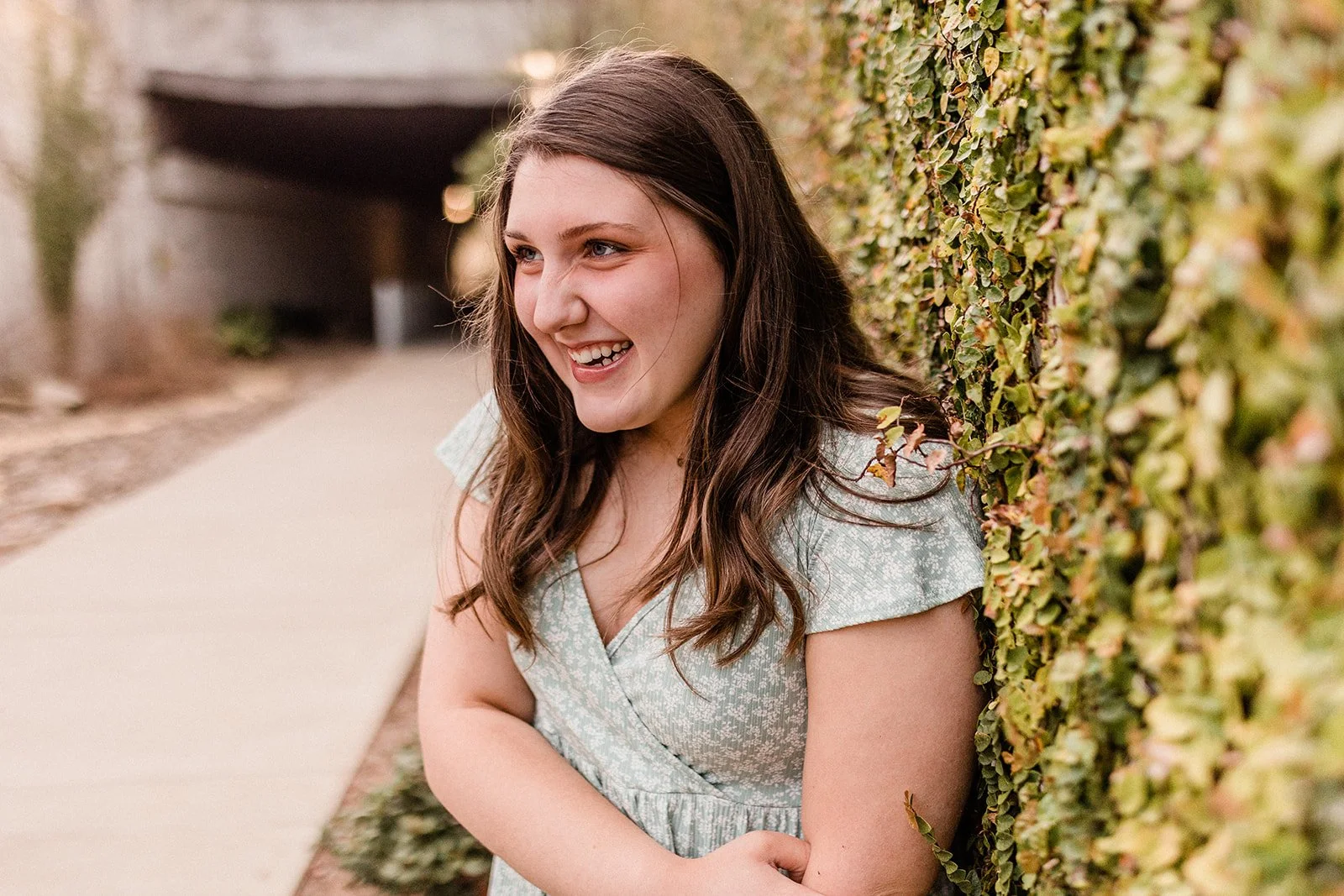 Railroad Park, Downtown Birmingham, AL - A young woman with long brown hair, wearing a light green patterned dress, laughing and leaning against a green leafy hedge outdoors.
