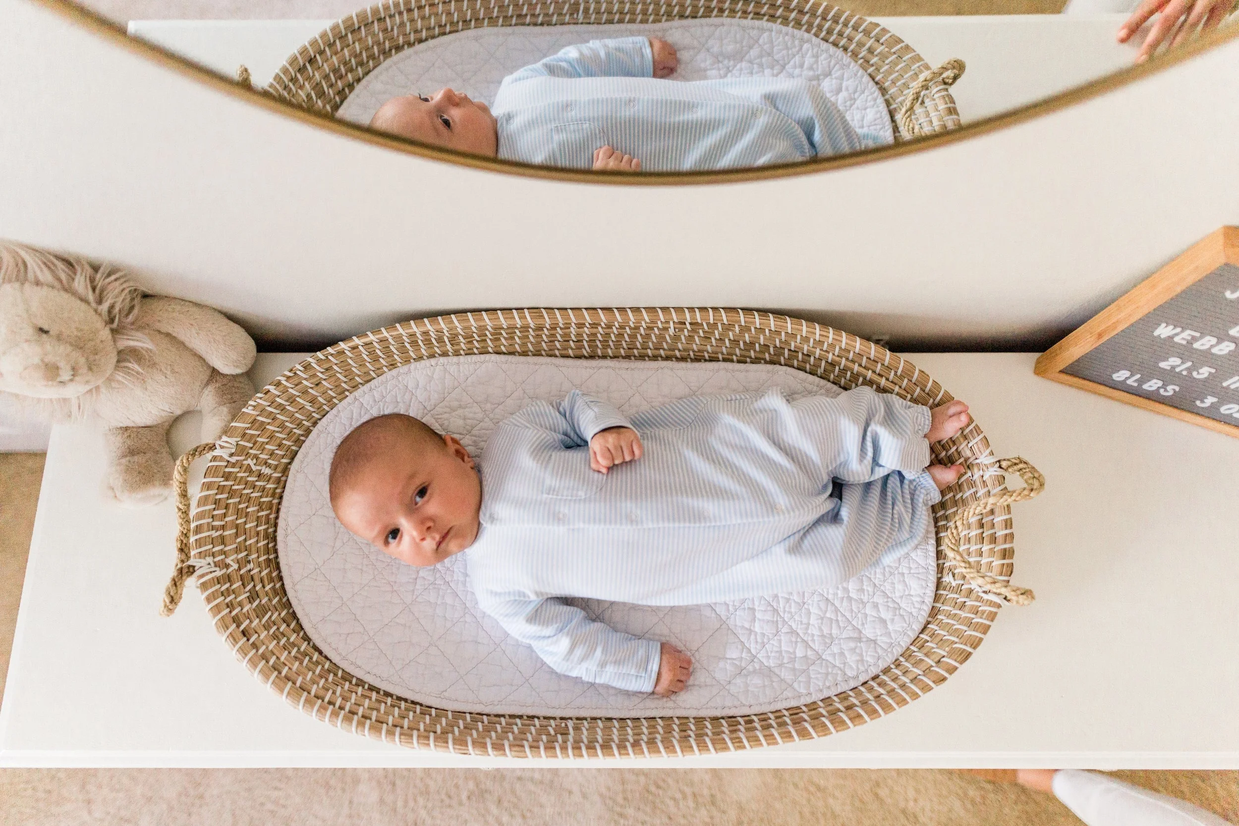Hoover, AL - A baby lying in a woven bassinet, wearing a light blue striped outfit, with a stuffed bunny and a mirror reflecting the baby.