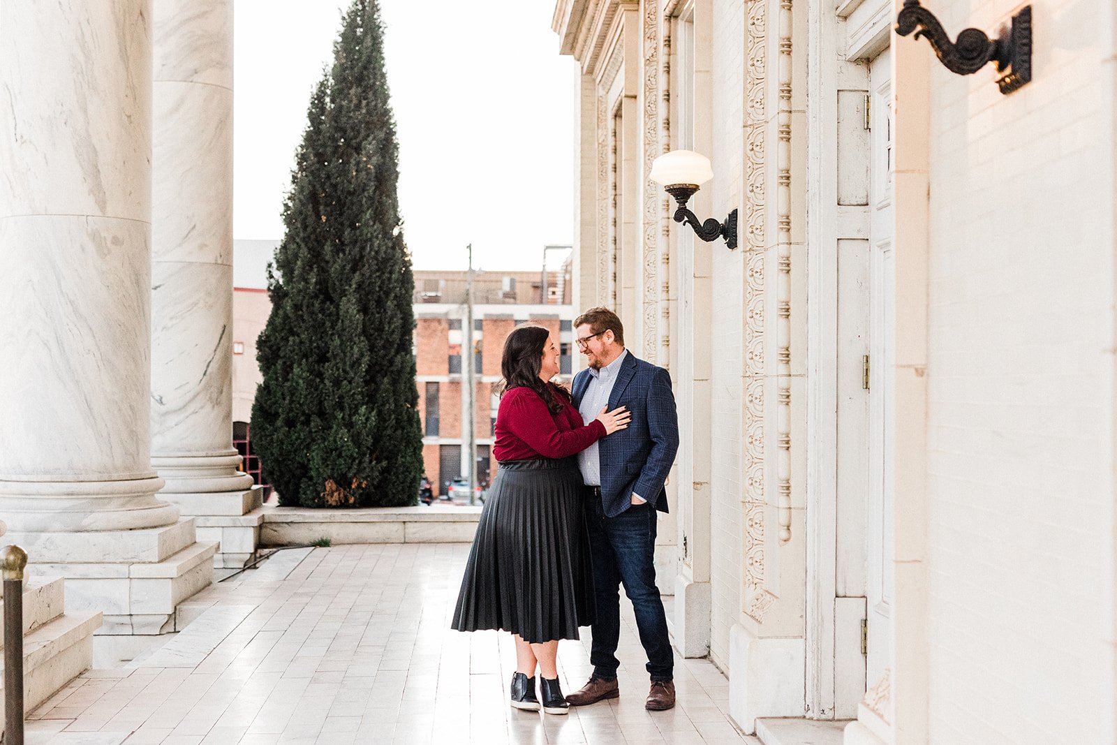 Downtown Birmingham, AL - A couple standing close together outdoors near a building with ornate white walls and black wall sconces, with a tall tree in the background. They are smiling at each other, the woman wearing a red top and black pleated skir