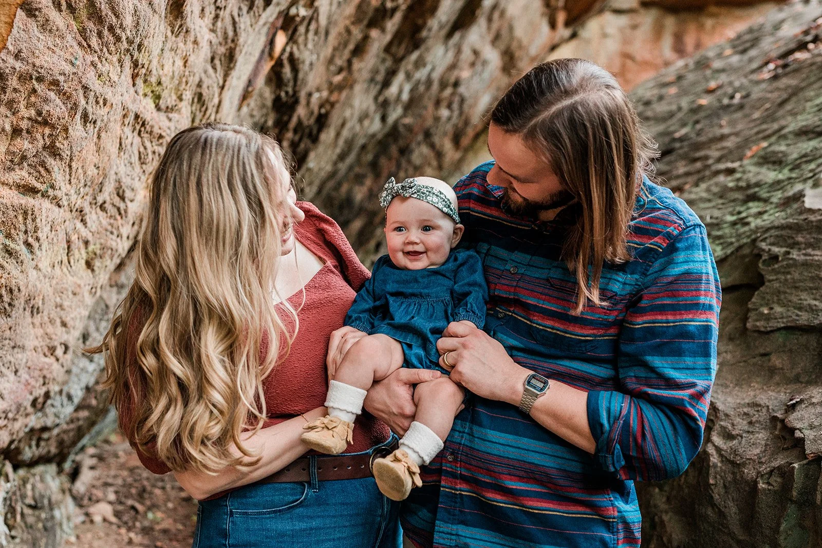 Moss Rock, Bluff Park, AL - A family of three enjoying time together outdoors inside a rocky area. The mother with long blonde hair wearing a red top is holding a baby girl with a bow headband and blue dress, while the father with long brown hair and