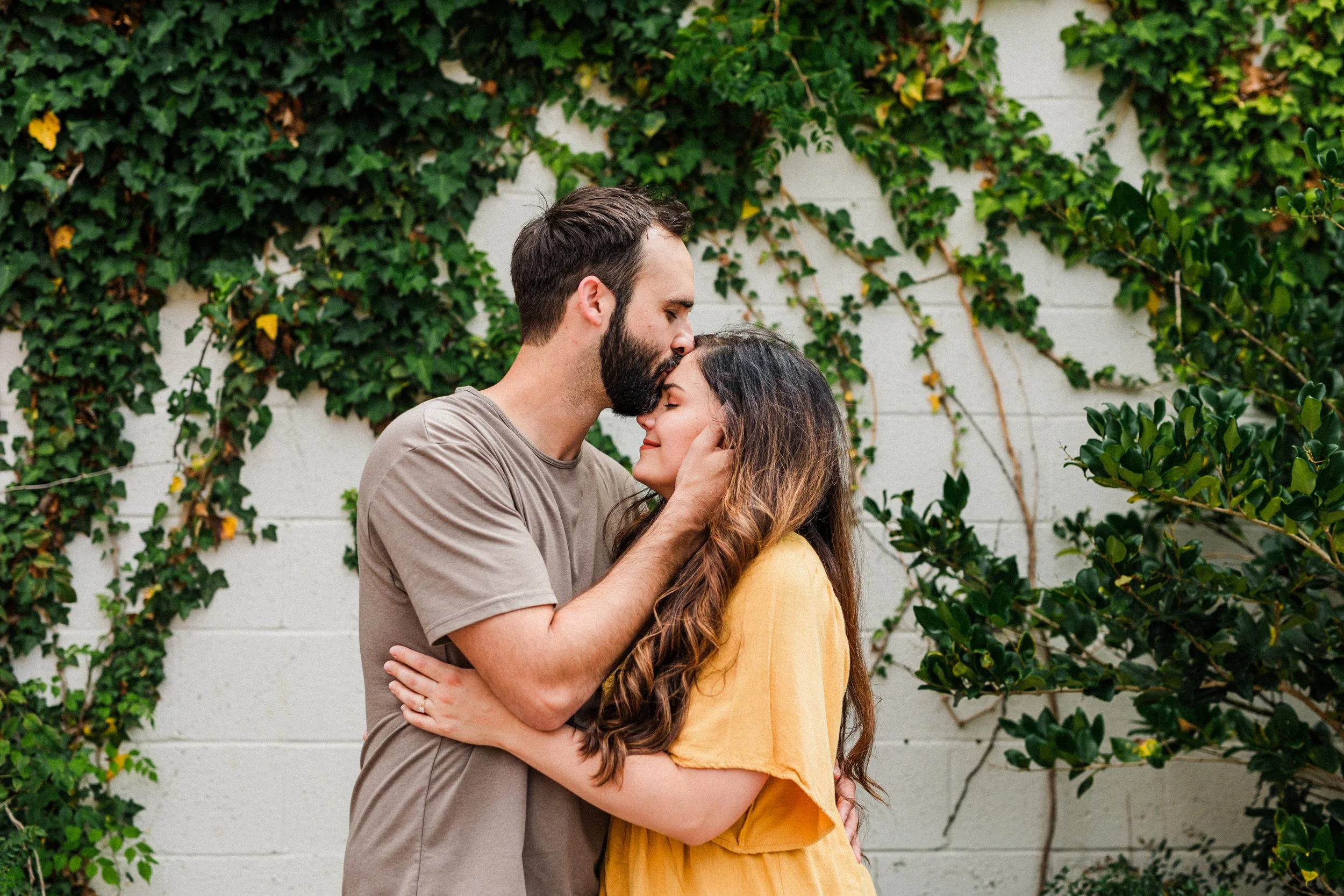 Opelika, AL - A man with a beard and a woman with long dark hair embrace and share a kiss against a background of green ivy and a white wall.