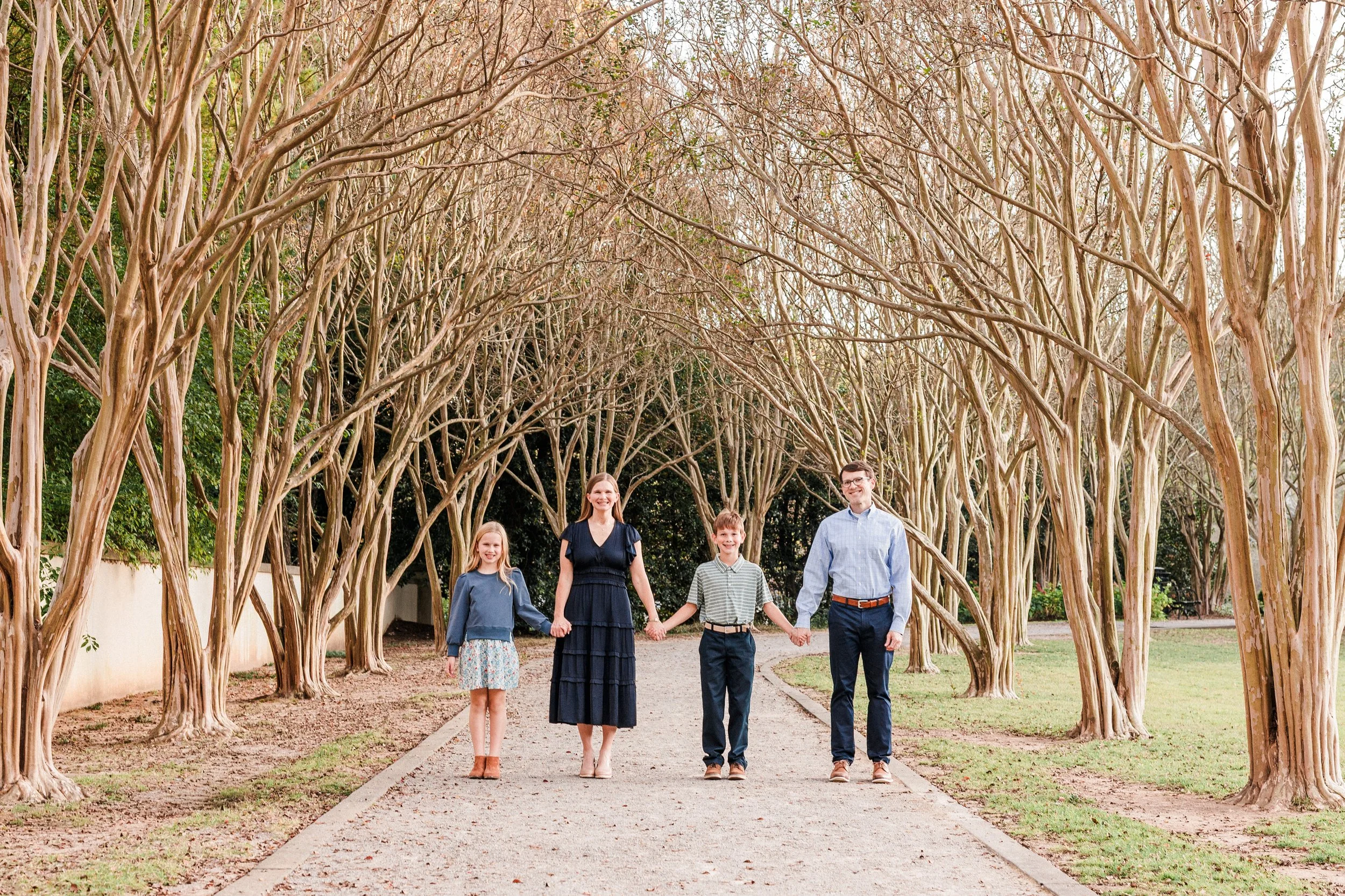 Birmingham, AL - A family of four walking hand-in-hand on a garden path under tall, leafless trees in a park during daytime.