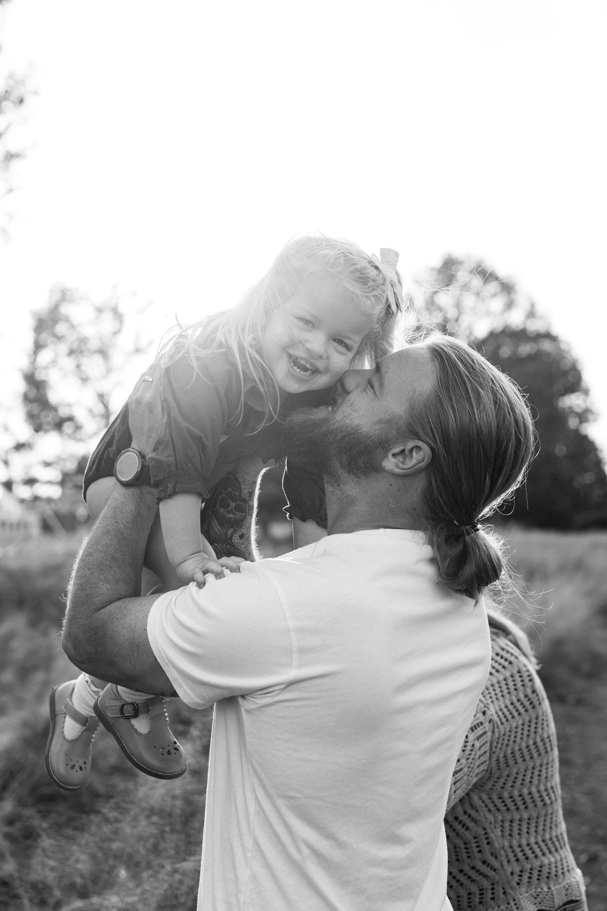 Railroad Park, Downtown Birmingham, AL - A man with long hair tied back, holding a young girl in a dress and sneakers, outdoors in a park, smiling and touching noses, with another woman partially visible in the background.