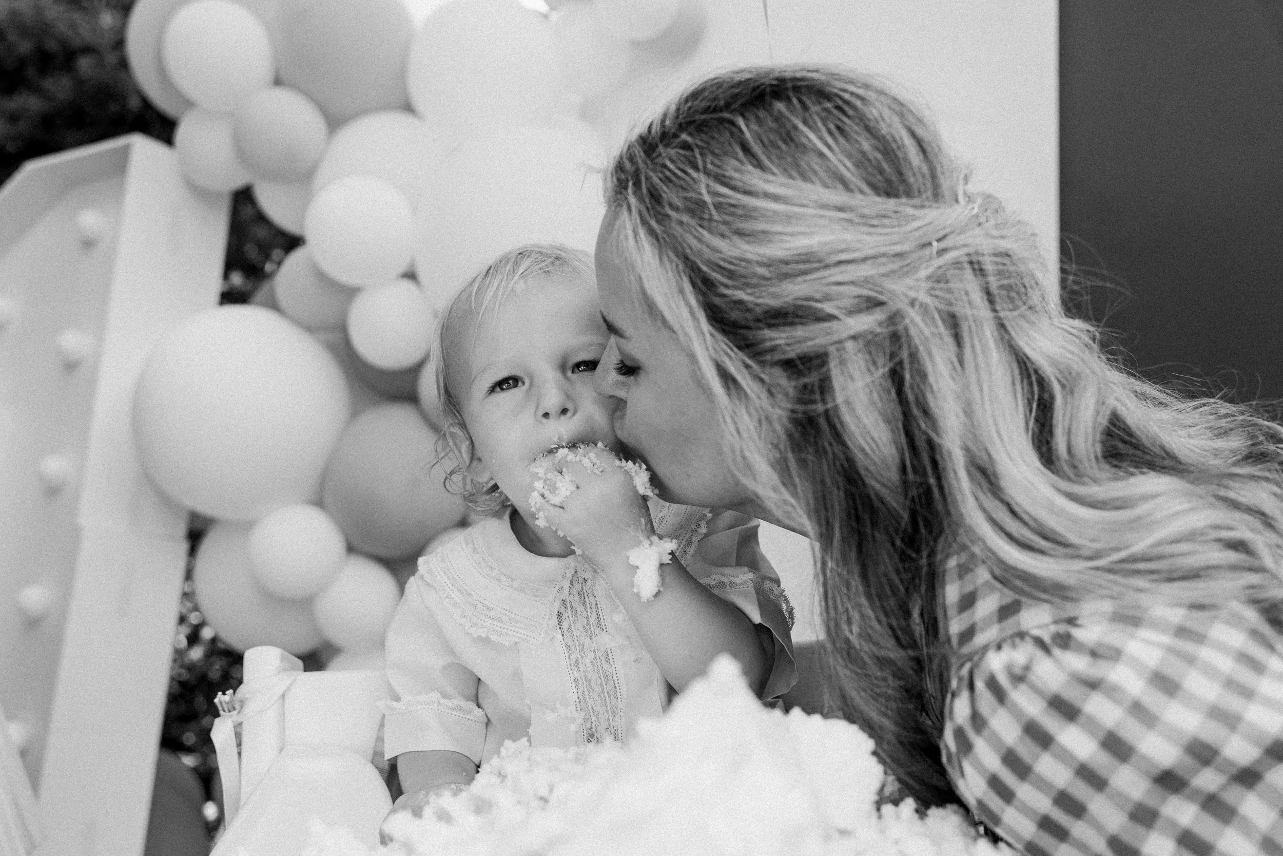 Mountain Brook, AL - A woman kisses a one year old on the cheek during a birthday celebration, with the boy eating cake and in front of balloons and a background.