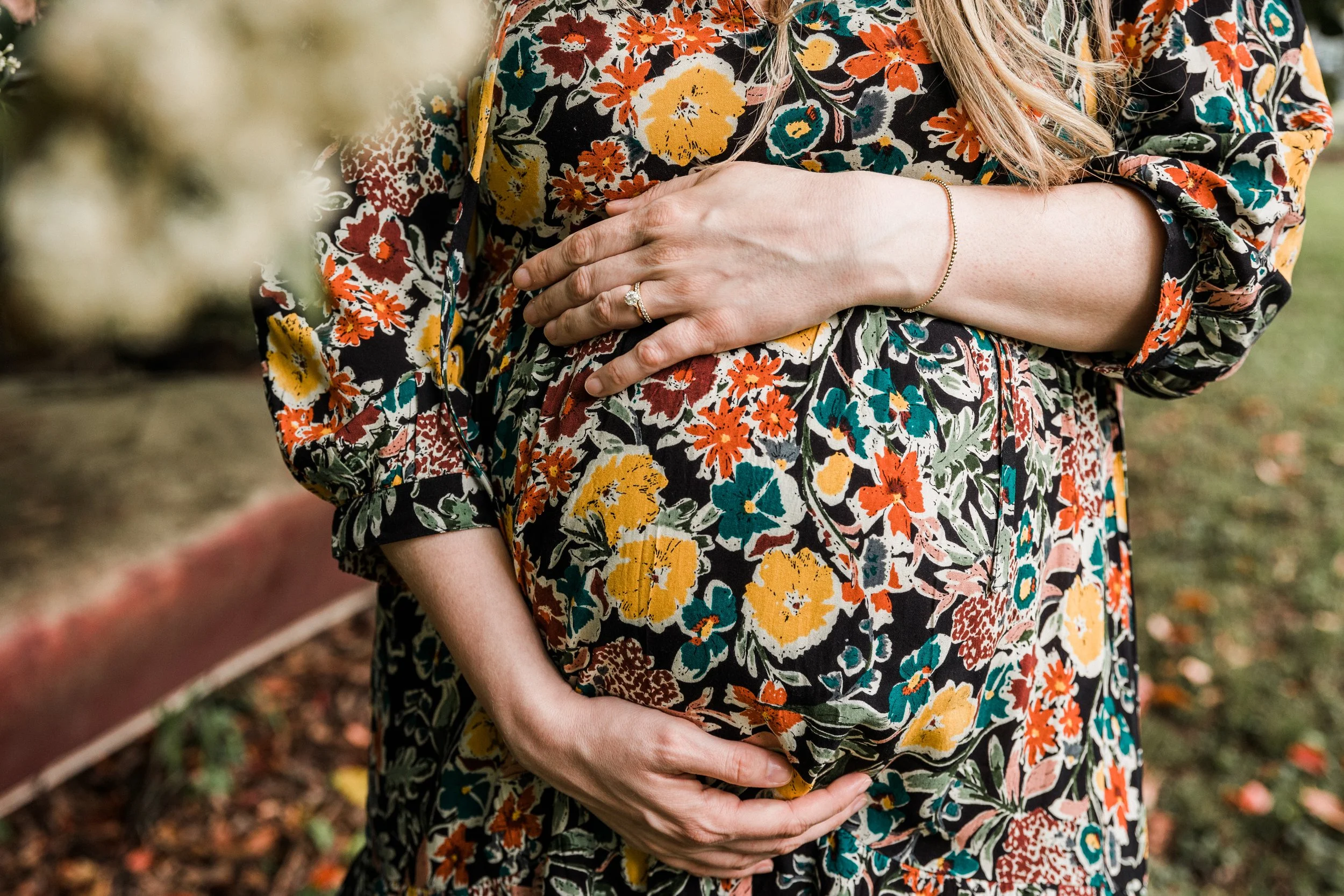 Vestavia, AL -Close-up of a pregnant woman wearing a floral dress, gently cradling her belly with both hands, outdoors with a blurred garden background.