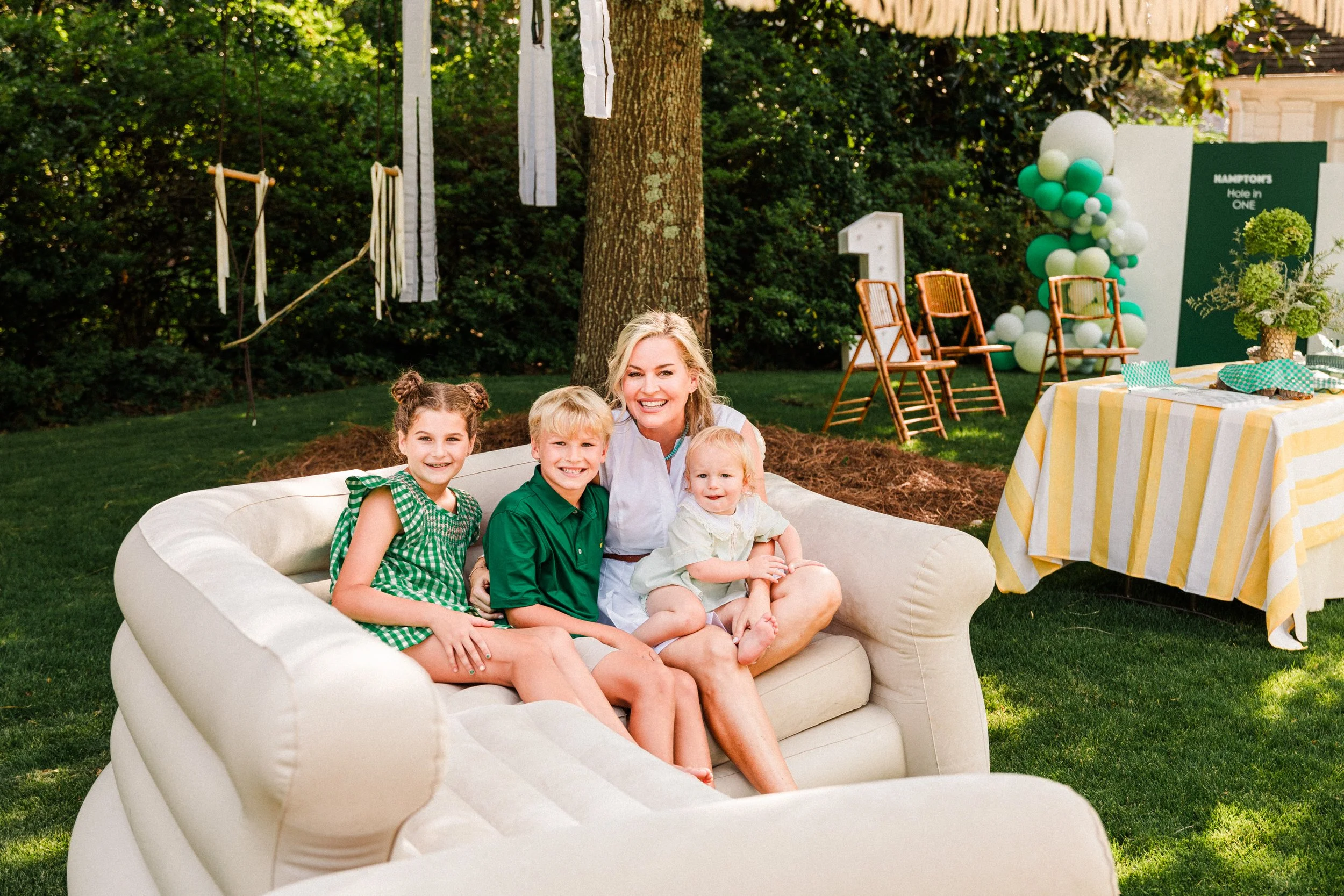 Mountain Brook, AL - A woman and three children sitting on a cream-colored couch outdoors during daytime, with a decorated setting in the background including balloons, chairs, and a table with a yellow and white striped tablecloth.
