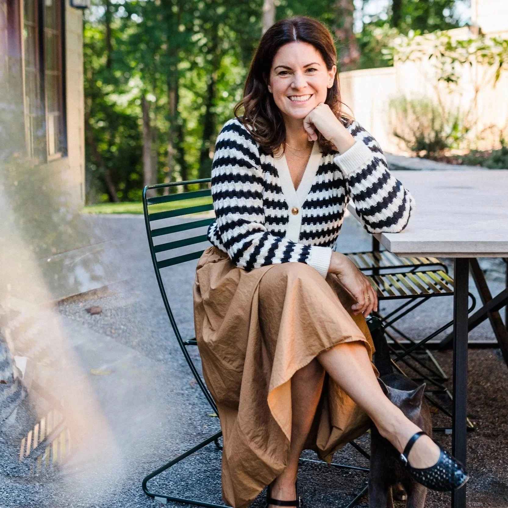 Cahaba Heights, AL - Woman sitting and smiling outdoors at a table with a cat walking beneath and a prism reflecting on the other side.