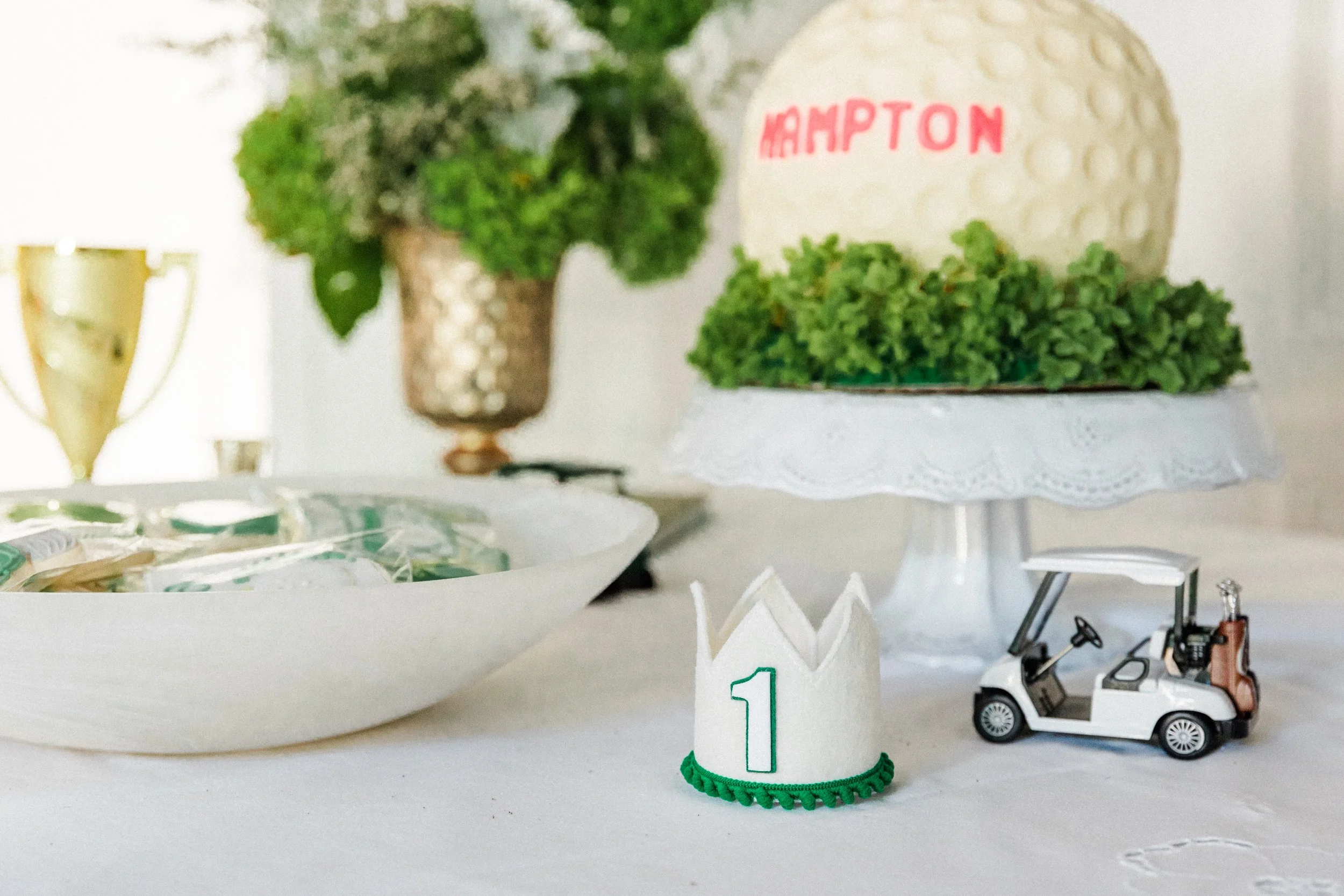 Mountain Brook, AL - Celebration table with a decorated cake, toy golf cart, a small crown with a number 1, a gold trophy, and a bowl of wrapped candies, indicating a first birthday party.