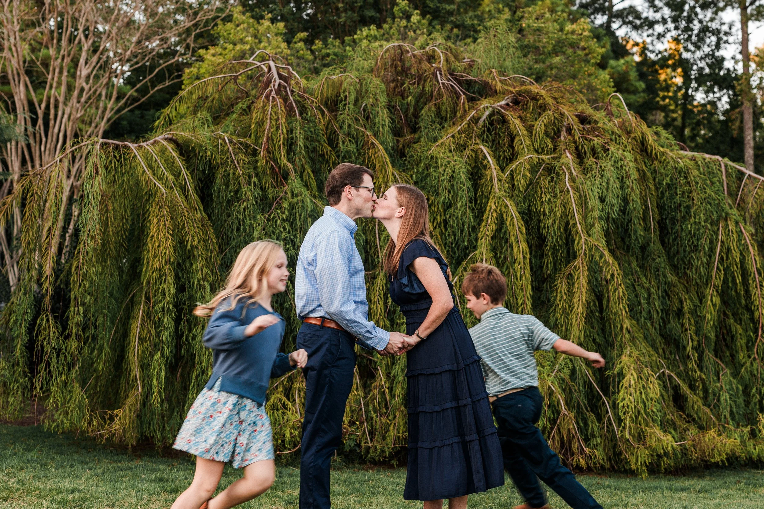 Birmingham Botanical Gardens - A couple shares a kiss while holding hands, surrounded by their children playing outdoors in front of a large, lush tree.