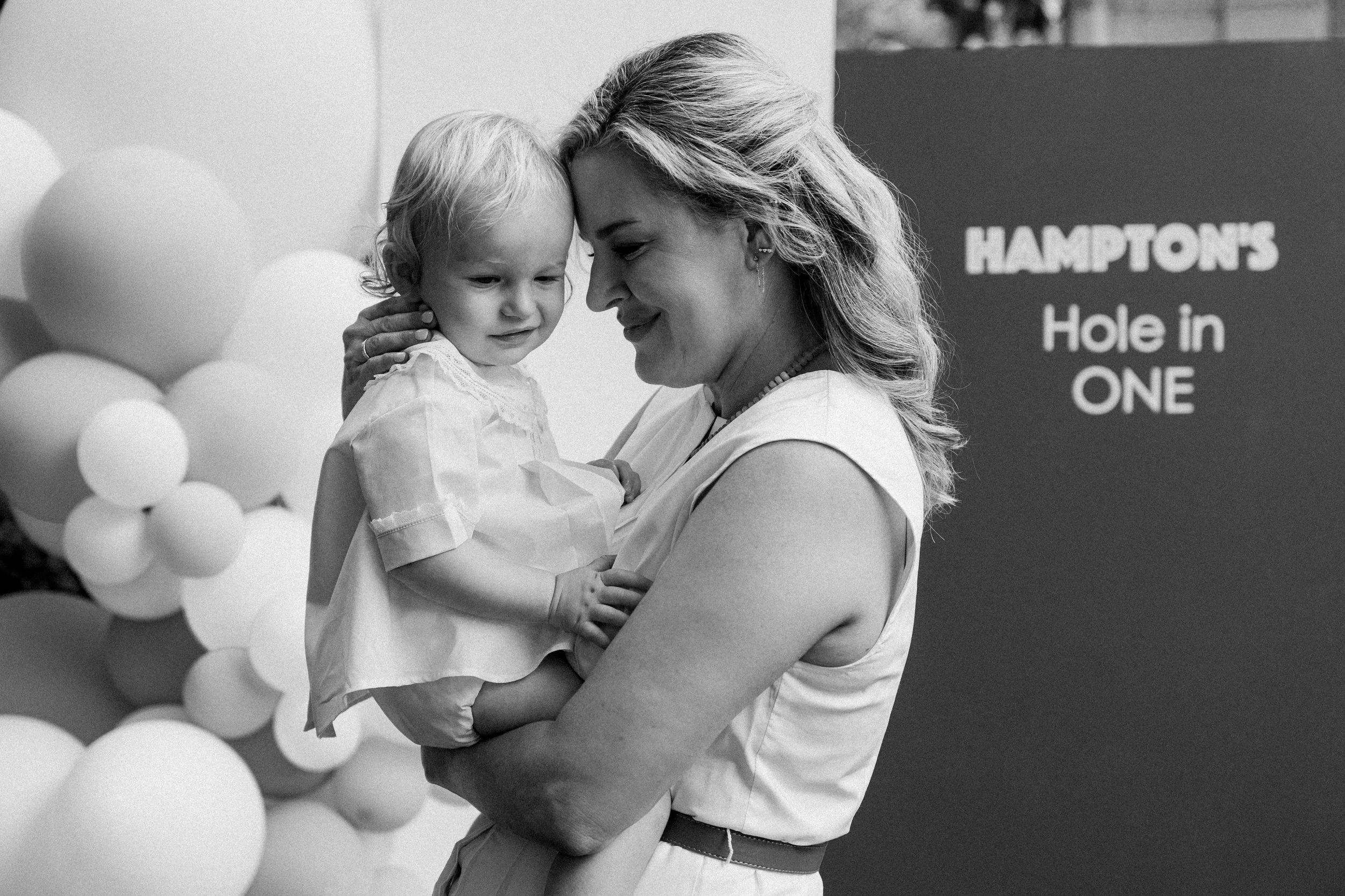 Mountain Brook, AL - A woman holding a young child in her arms, both with closed foreheads touching, smiling gently. Background includes balloons and a sign that says 'HAMPTON'S Hole in ONE.'