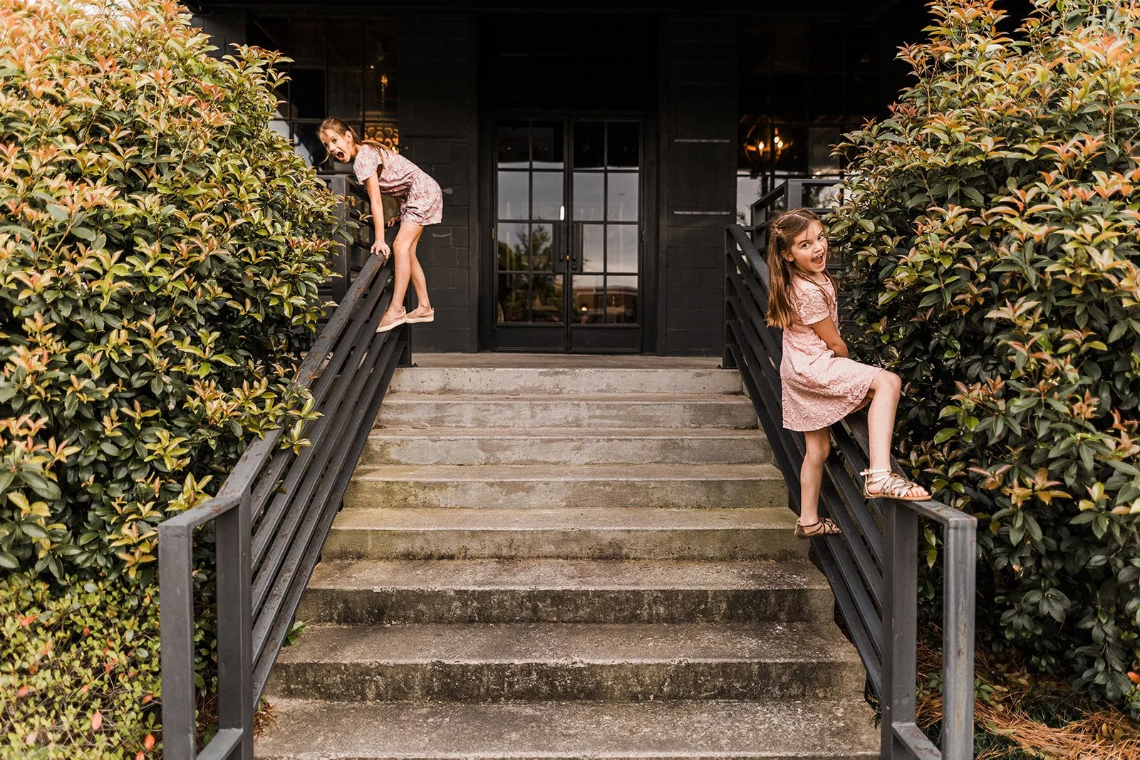 Pepper Place, Downtown Birmingham, AL - Two young girls in pink dresses playing on the railings of a staircase outside a black building with large windows, surrounded by bushes.