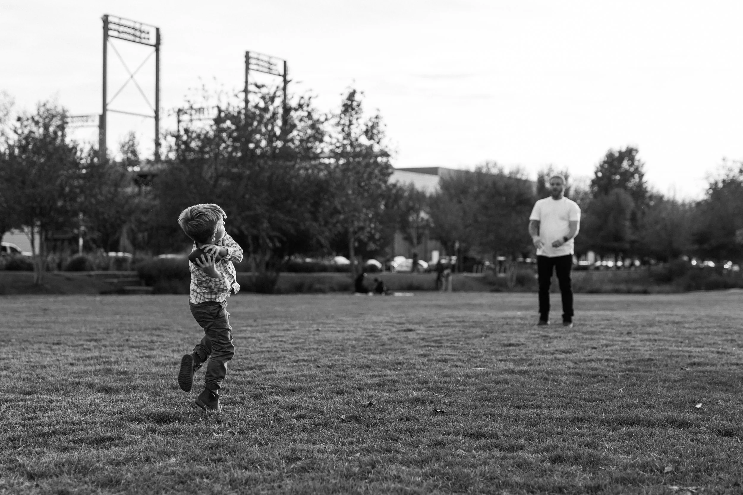 Railroad Park, Downtown Birmingham, AL - A young boy in a checkered shirt running on a grassy field with a football, while an adult man stands in the background watching, in a park with trees and structures in the distance, in black and white.