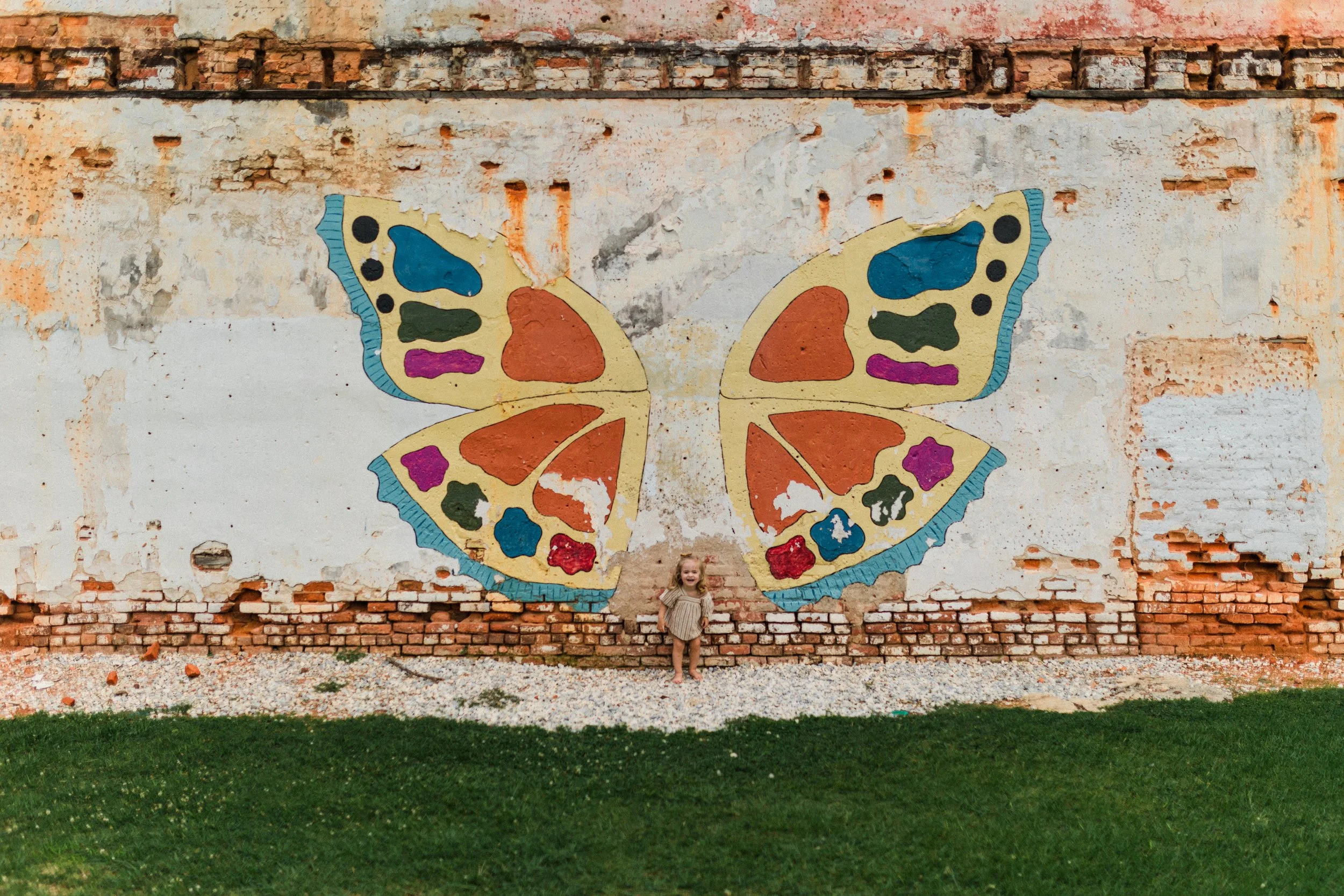 Opelika, AL - A young girl standing in front of a wall painted with a large, colorful butterfly mural. The wall has a weathered, partially exposed brick and plaster surface, and the ground is a mix of gravel and grass.