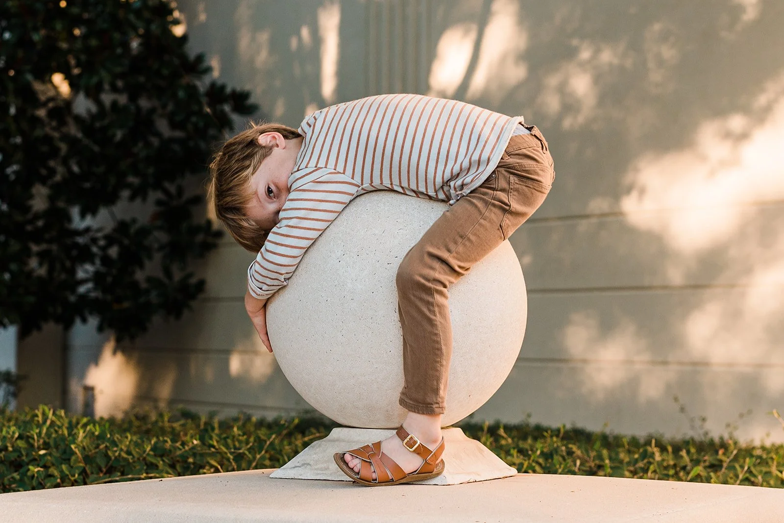 Vestavia, AL -A boy with light brown hair wearing a striped shirt and brown pants, lying on top of a large rounded stone sculpture in an outdoor setting during late afternoon or early evening.