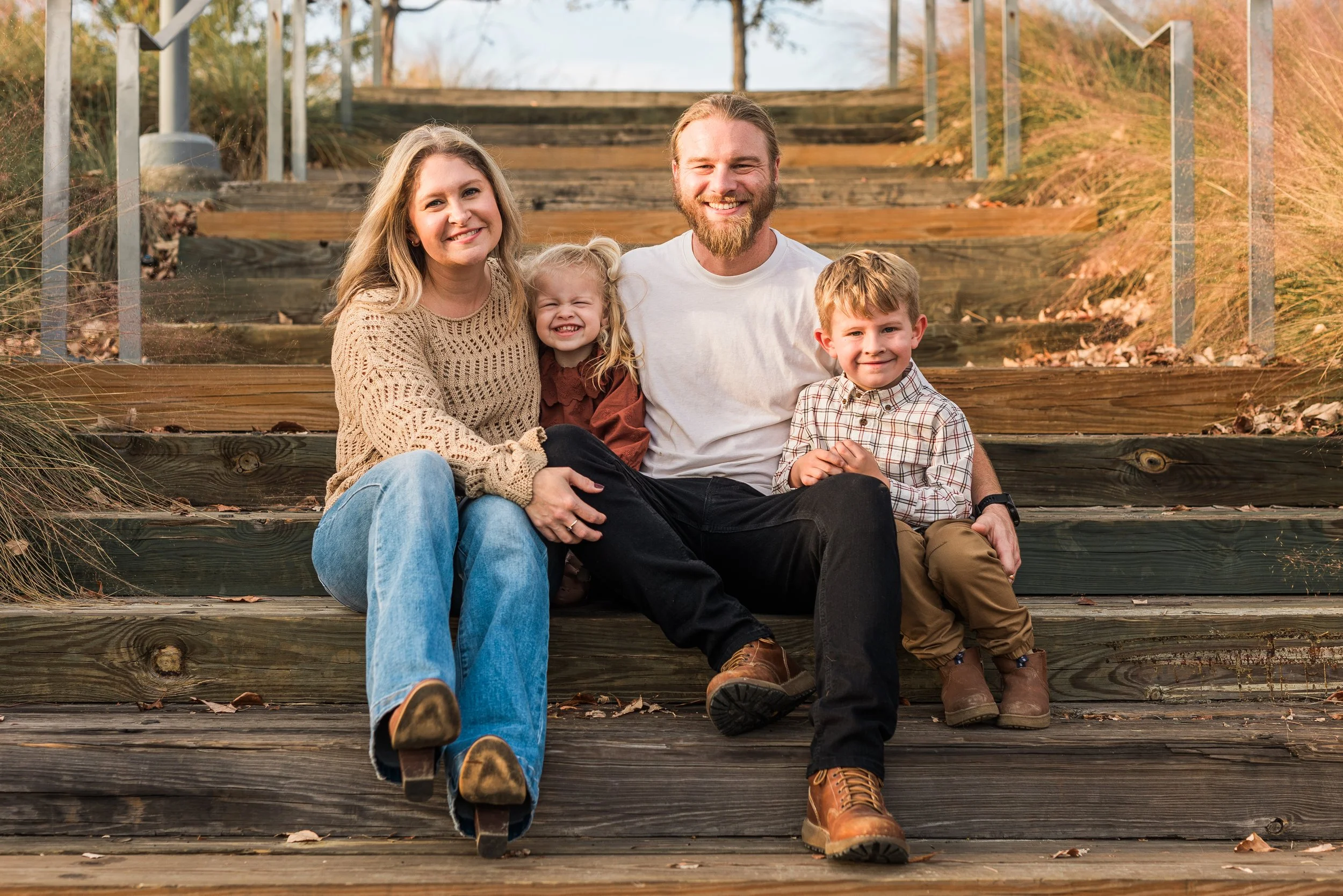 Railroad Park, DowntA family of five sitting on wooden stairs outdoors, smiling at the camera. The family includes a woman, a man, a young girl, a young boy, and a toddler girl. The background features natural landscape with grass and autumn foliage.