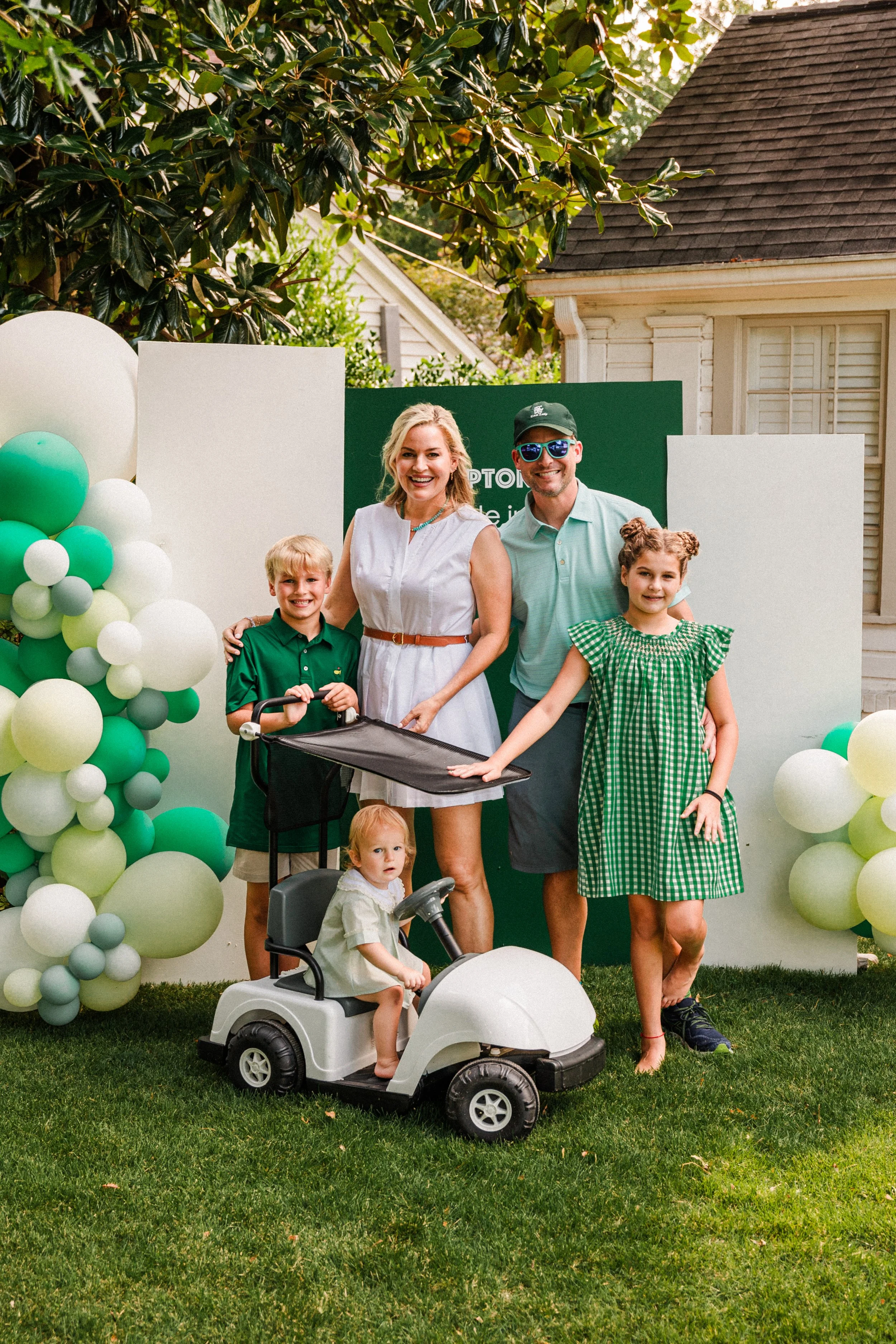 Mountain Brook, AL - Family gathering outdoors with balloons and a green backdrop, including a woman, man, and three children, one of whom is on a toy car, smiling for a photo.