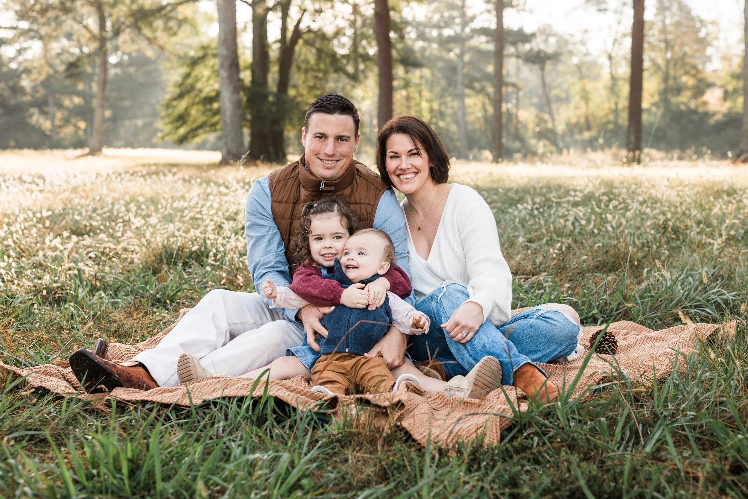 Vestavia, AL -A happy family of four sitting on a blanket in a sunlit park surrounded by tall trees, smiling at the camera.