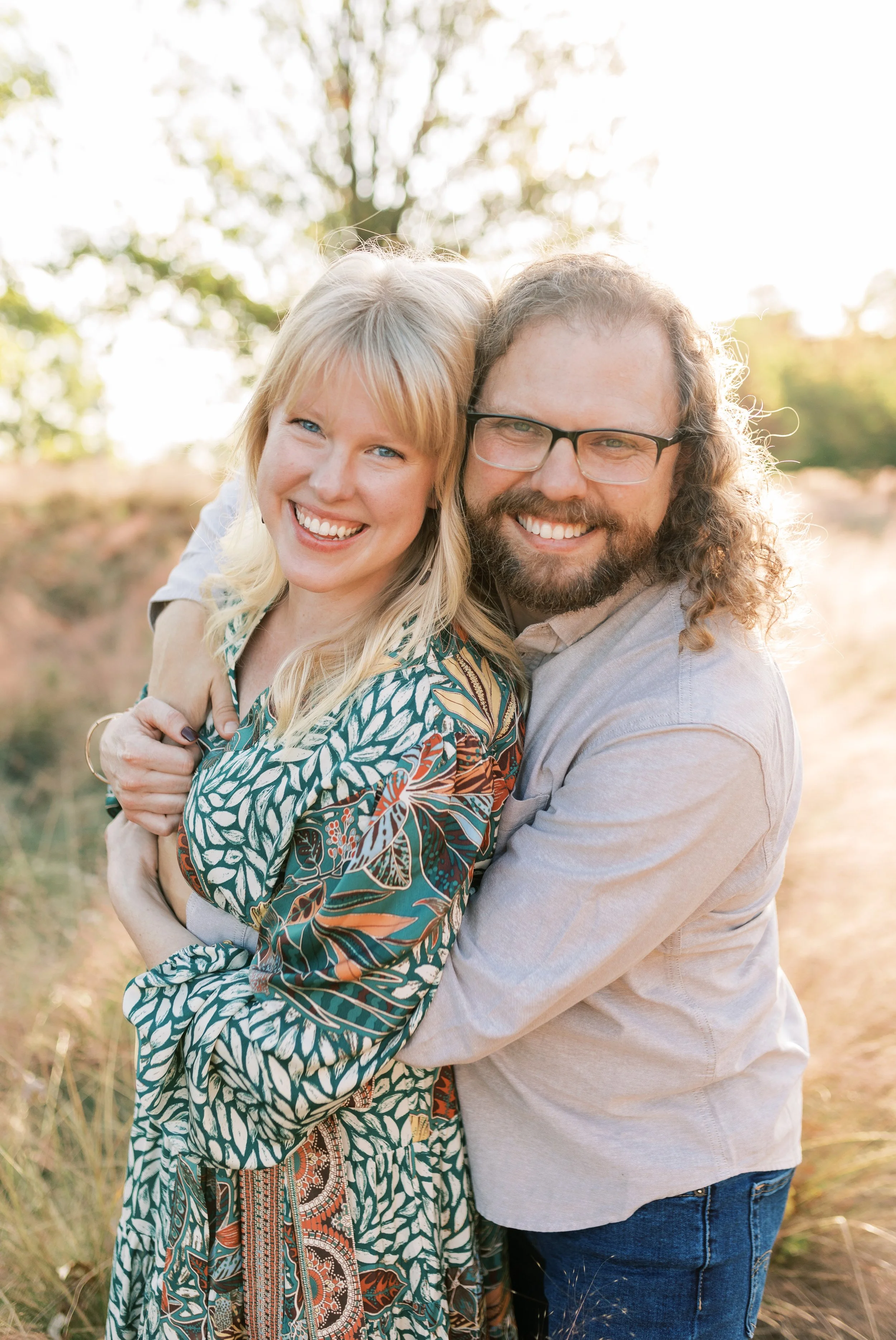 Railroad Park, Birmingham, AL - A smiling couple embracing outdoors in natural light, with trees and a dirt path in the background.