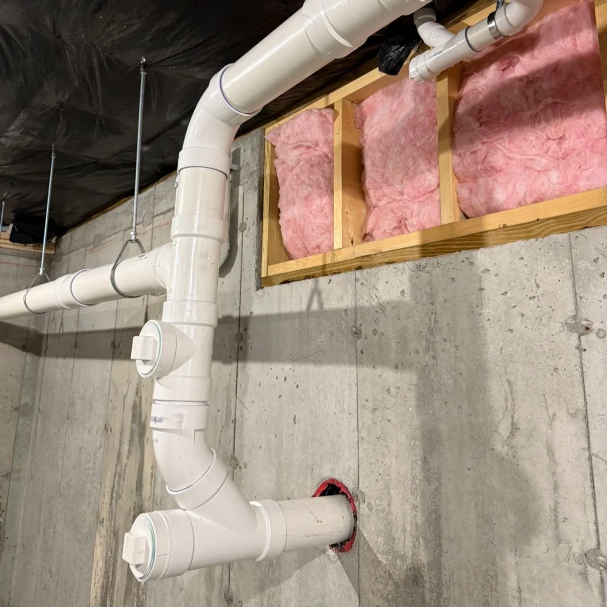 Basement or utility room with exposed concrete walls and floor. White PVC pipes running along the wall and ceiling, connected with various joints and fittings. Insulation pink fiberglass batts stored in a wooden frame mounted on the wall.