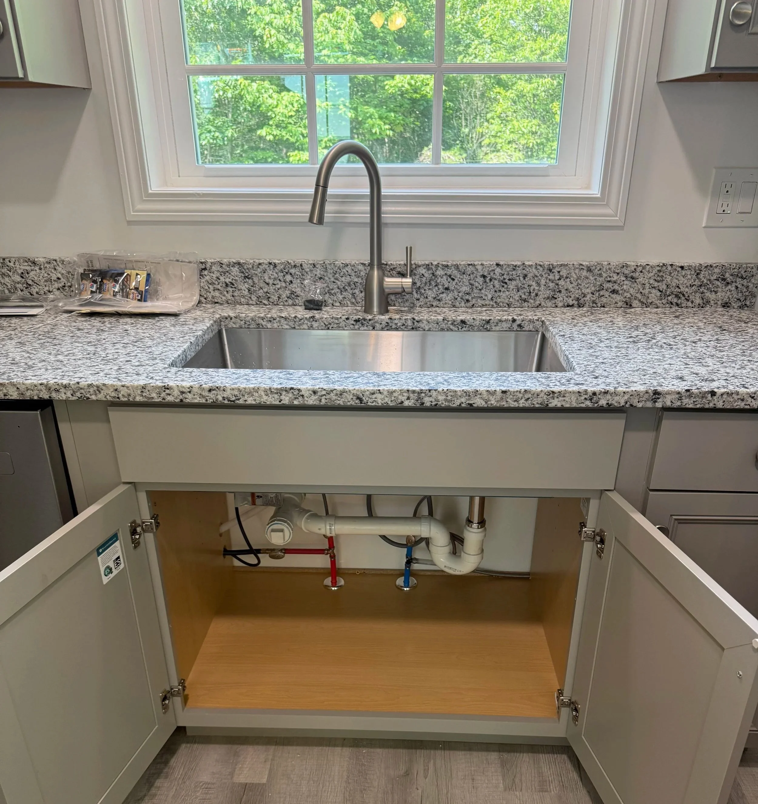 Kitchen sink under a window with some plumbing exposed beneath the cabinet. The countertop is granite, and there are cabinets on either side.